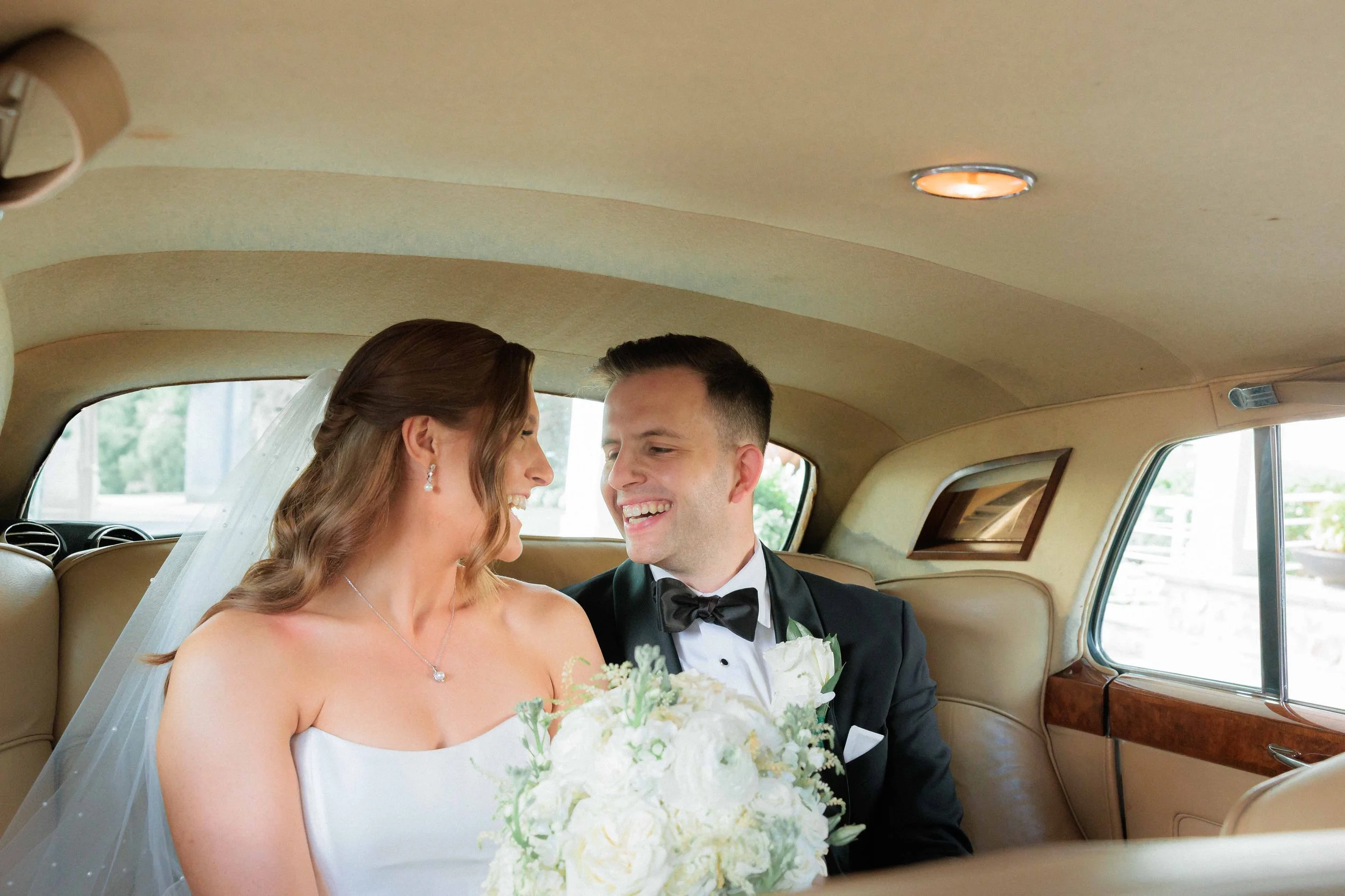 Bride and groom smiling and looking at each other inside a vintage car, bride holding a bouquet of white flowers, wedding attire, wedding celebration.
