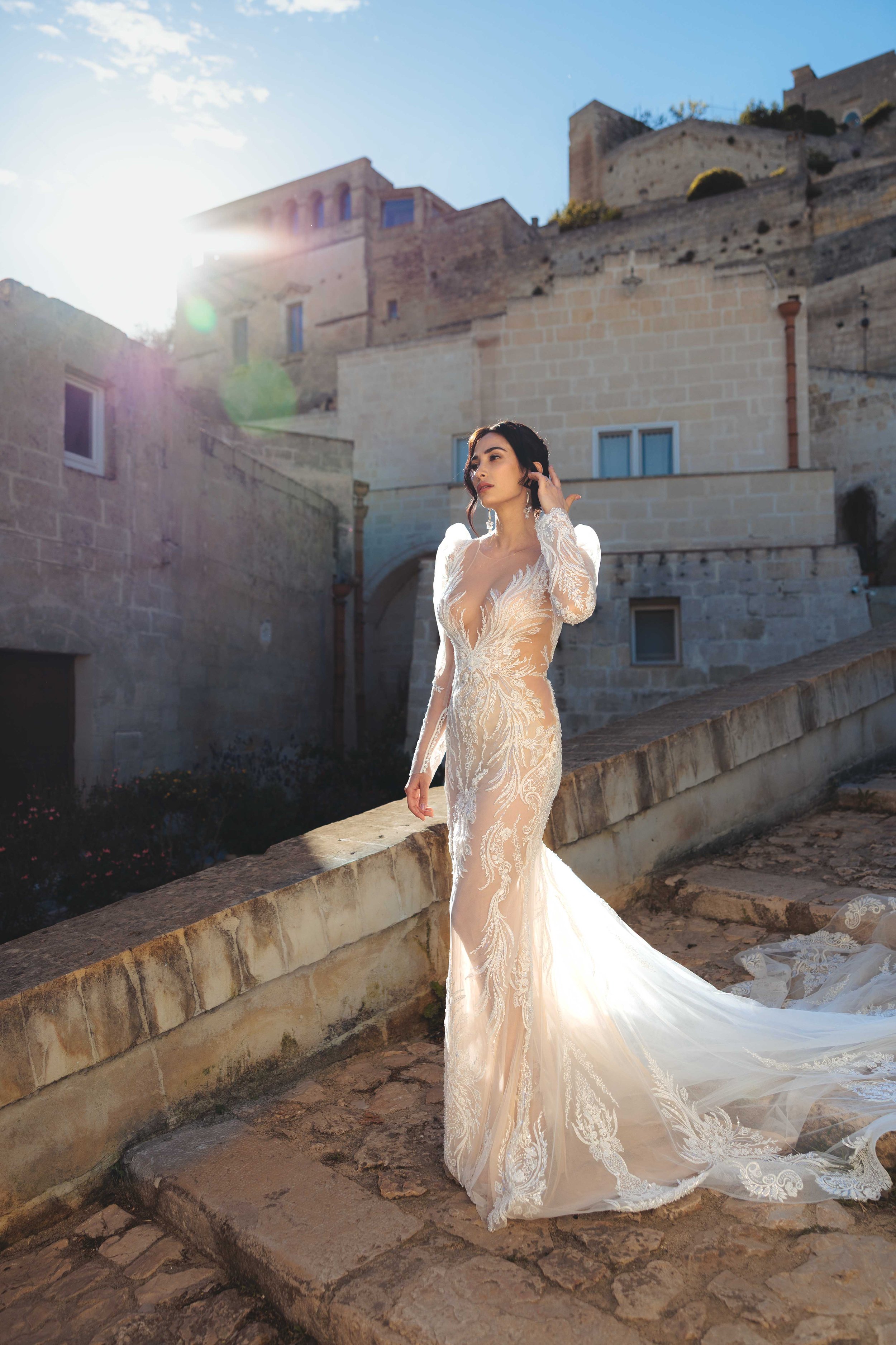 A woman in a white wedding gown standing outdoors on a cobblestone path, with historic stone buildings and a bright sun in the background.