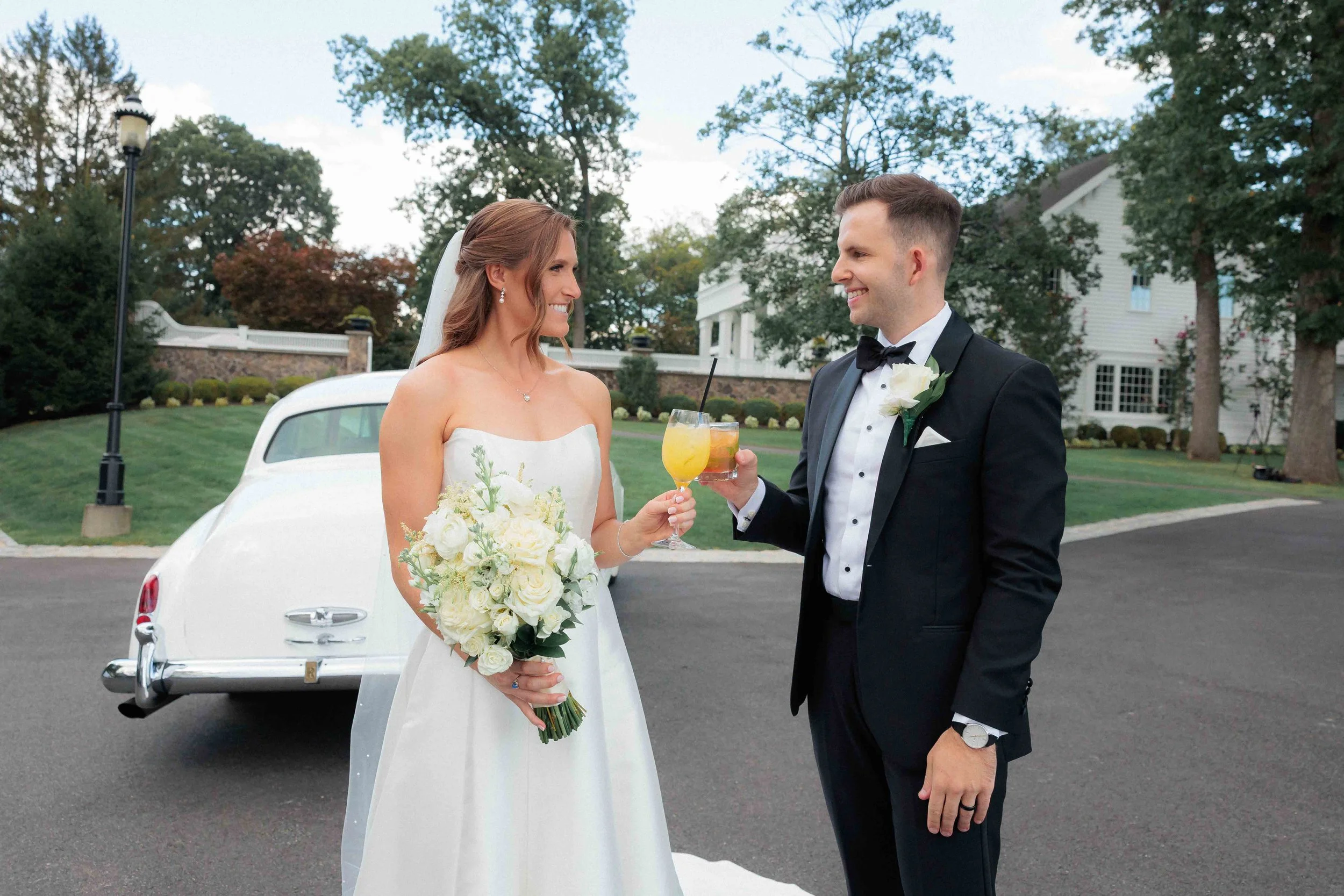 Bride and groom celebrating with drinks in front of a classic white car on their wedding day, outdoor setting with green trees and a large white house in the background.