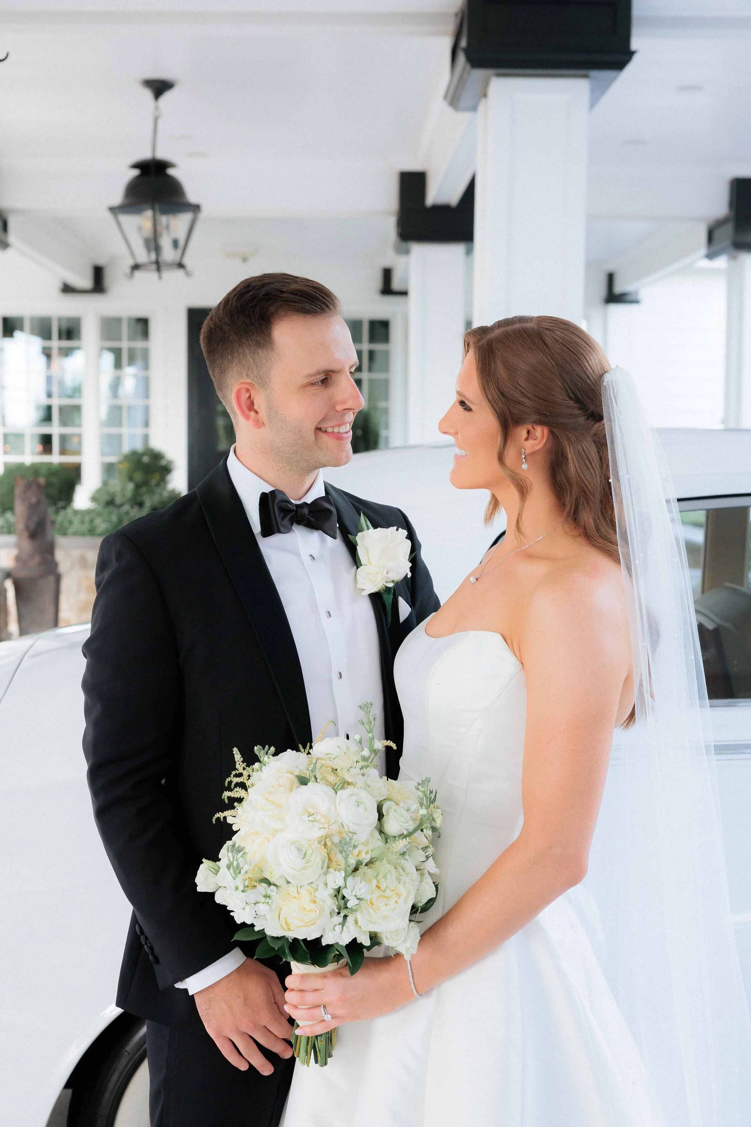 A bride and groom smiling at each other in wedding attire, standing in front of a white car with a house in the background.