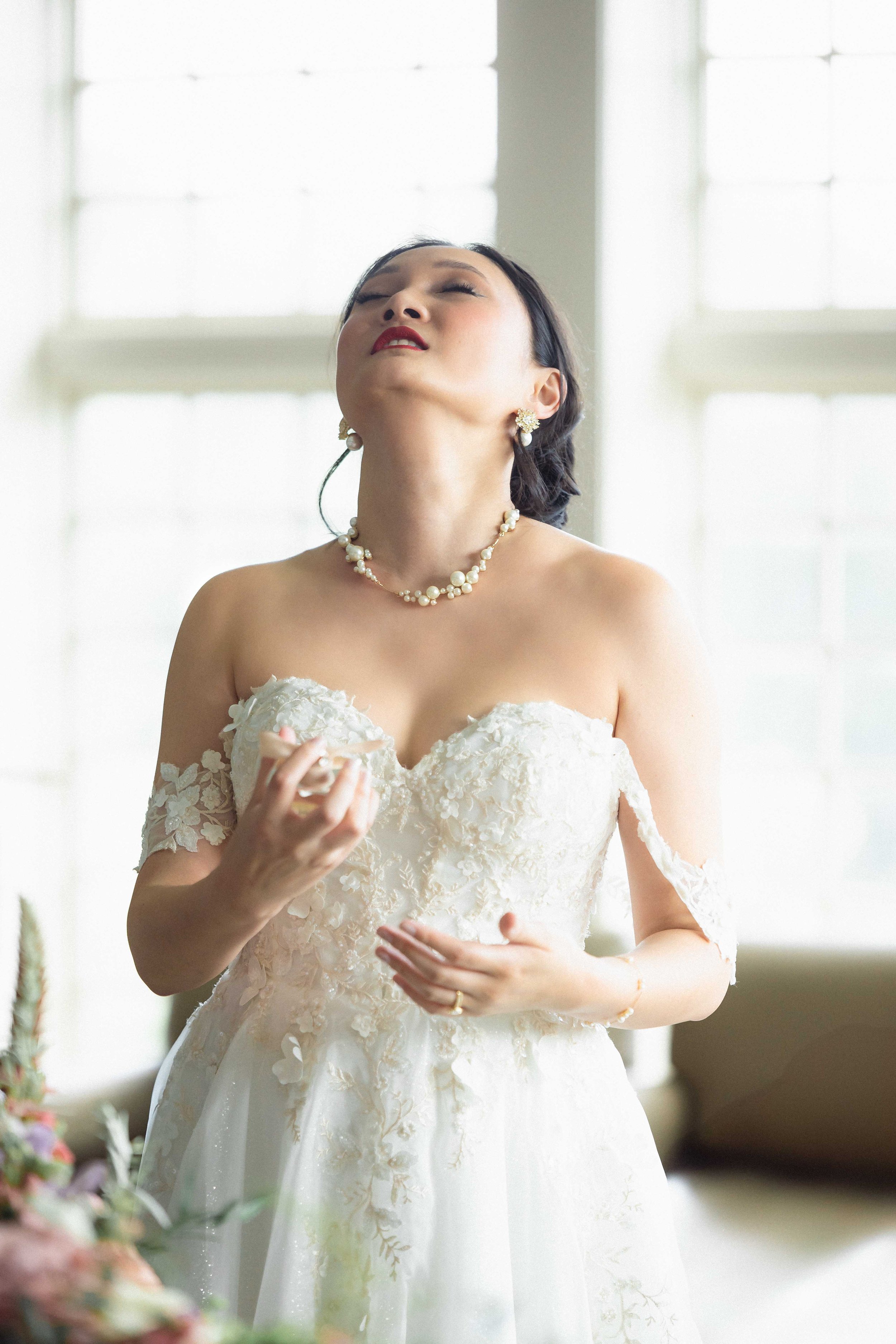 Brideswoman in an off-shoulder wedding dress with floral lace details, wearing pearl jewelry, standing indoors with closed windows in the background.