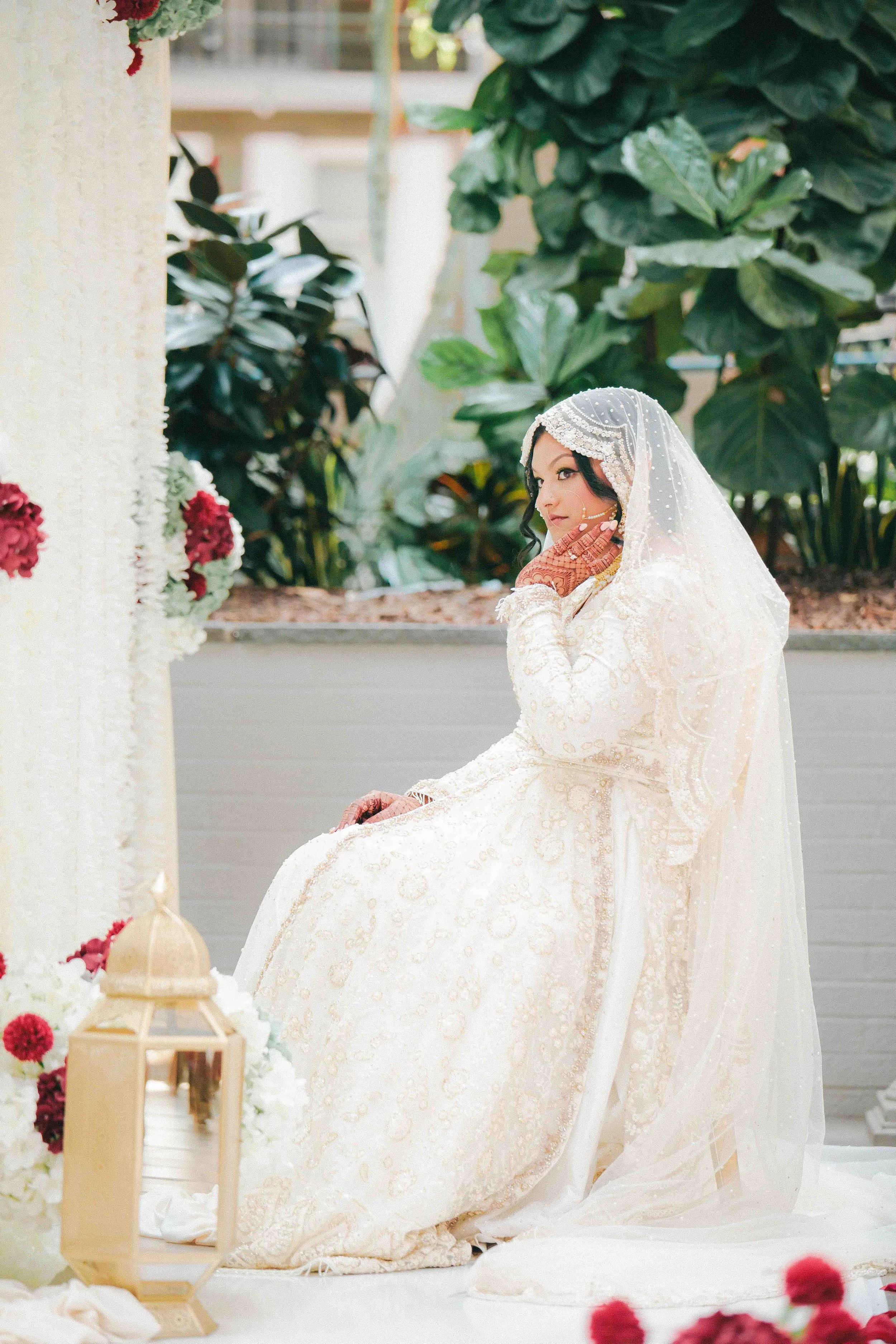 A bride in a white wedding dress with detailed embroidery, wearing a veil and henna on her hands, seated indoors in front of a floral and greenery backdrop.