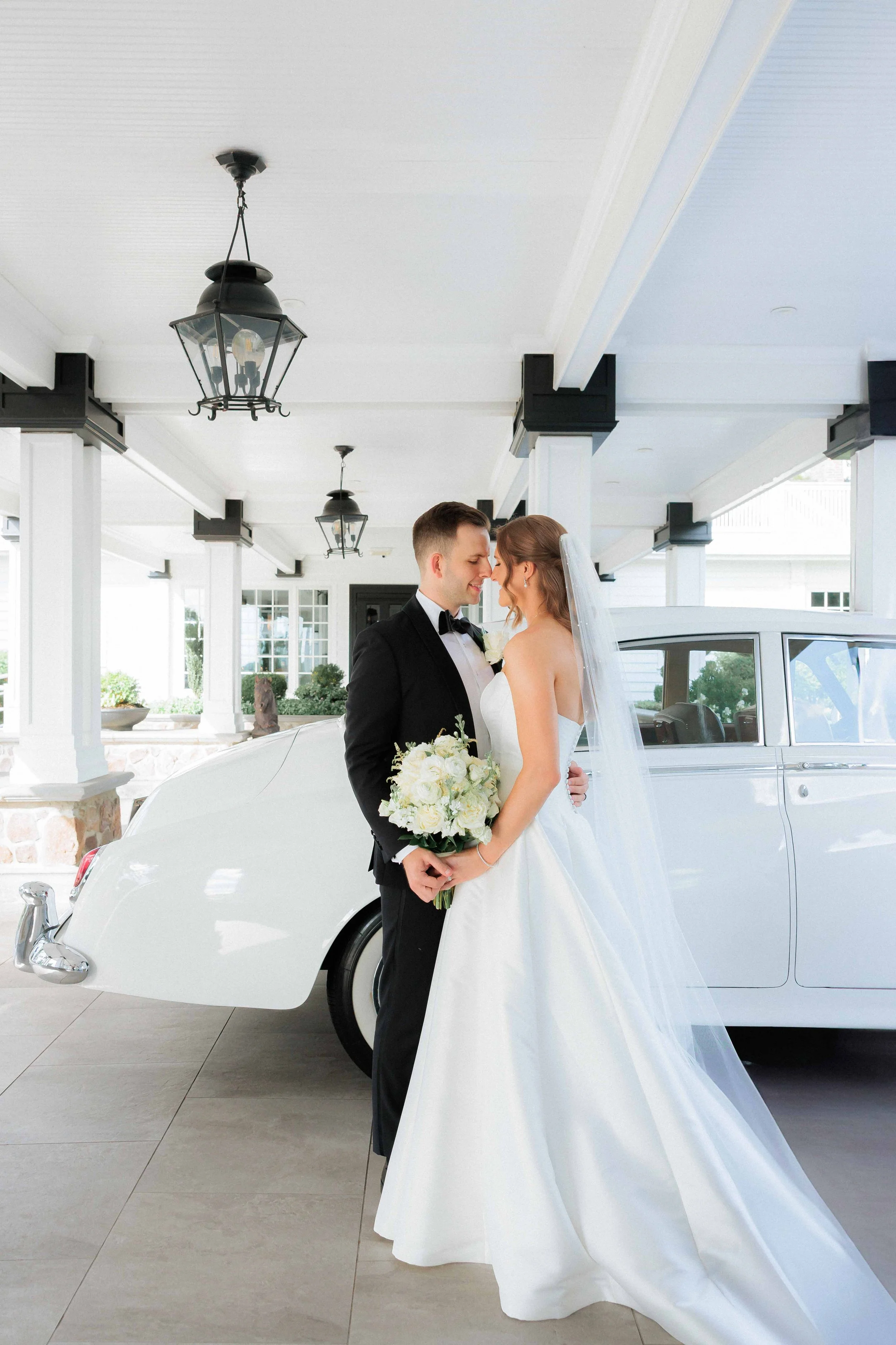 Bride and groom smiling and touching foreheads in front of a classic white car at a wedding.