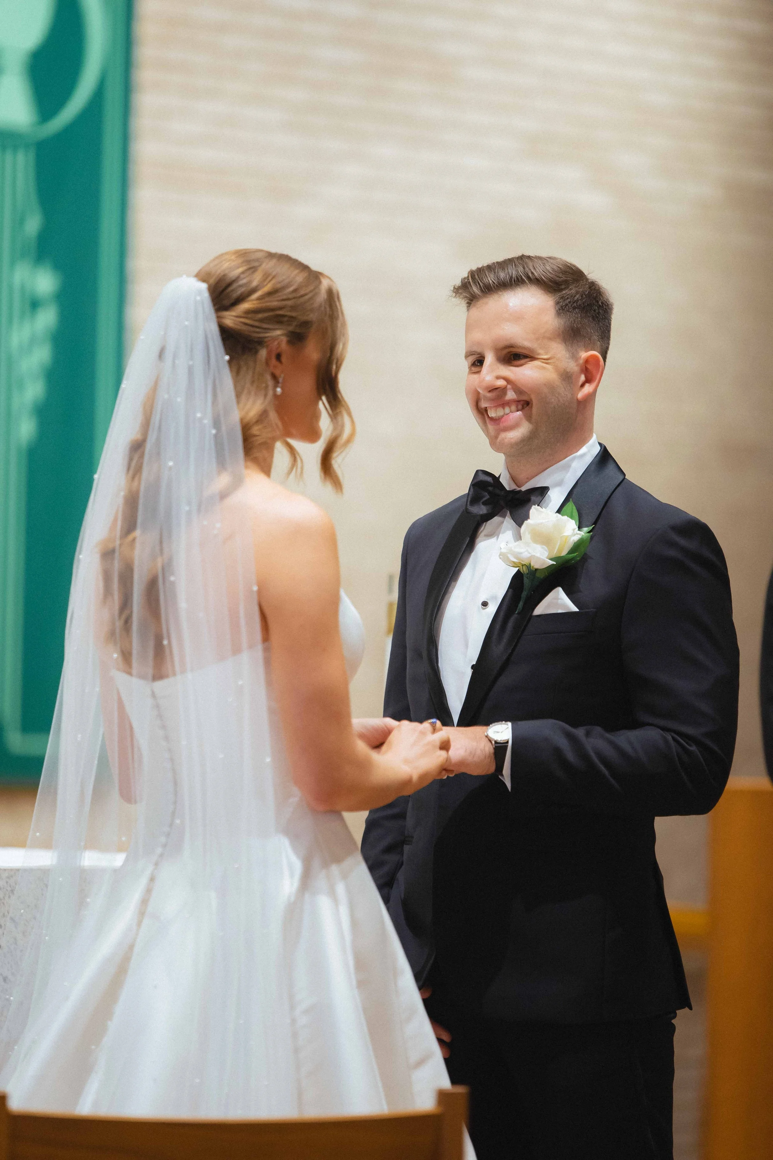 A bride and groom exchanging vows during their wedding ceremony. The bride wears a white wedding gown and veil, and the groom is dressed in a black tuxedo with a white shirt, black bow tie, and a white flower boutonniere. They are holding hands and s