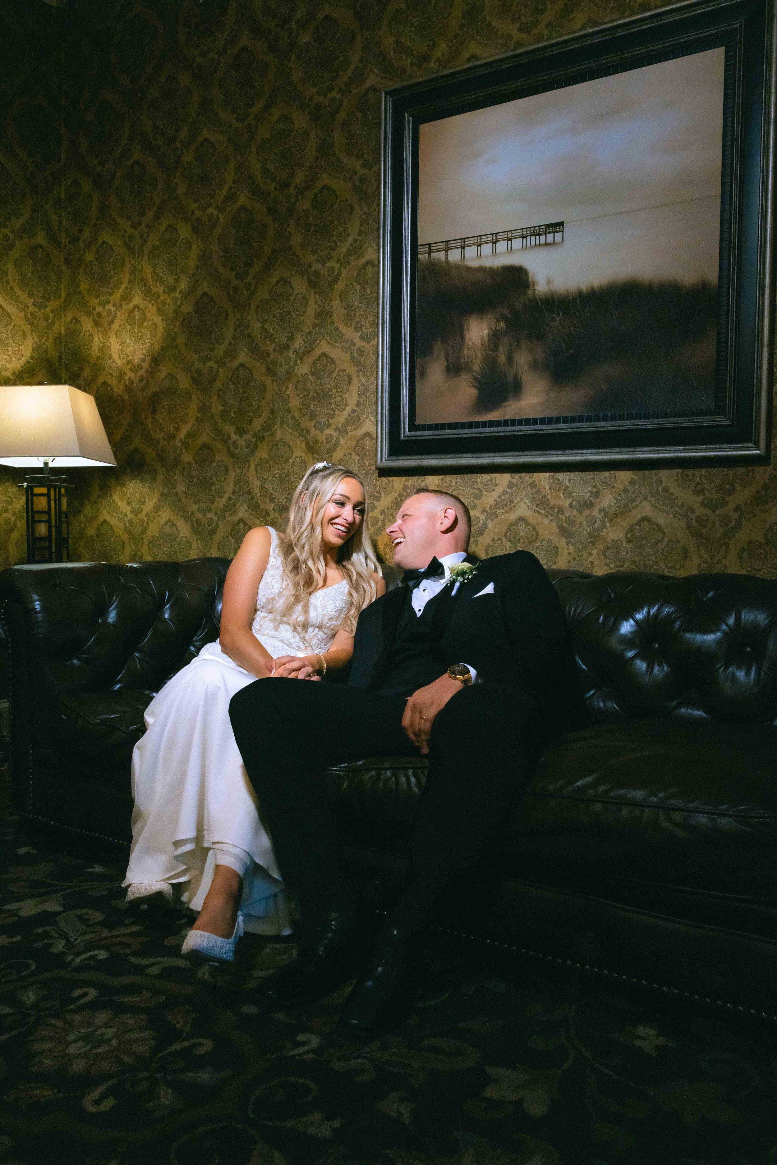 A bride and groom sitting on a black leather couch inside The Old Daley on Crooked Lake wedding venue, smiling and looking at each other, with a framed painting of a pier over water hanging on a patterned wallpapered wall behind them.
