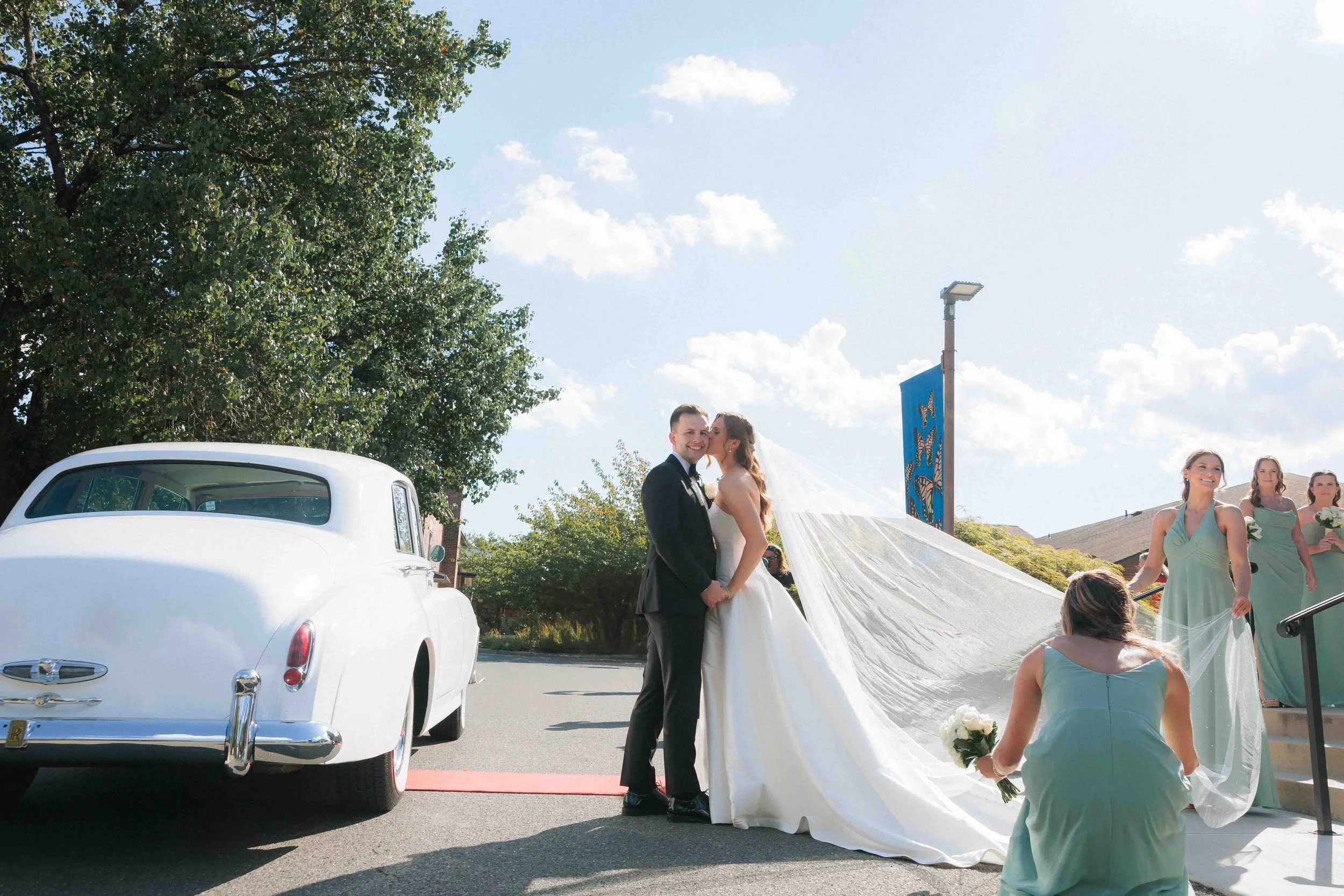 A bride and groom on their wedding day, with the bride wearing a long white dress and veil, standing next to a vintage white car. Bridesmaids in matching pastel green dresses are near the steps, holding bouquets, with one kneeling in front of the cou