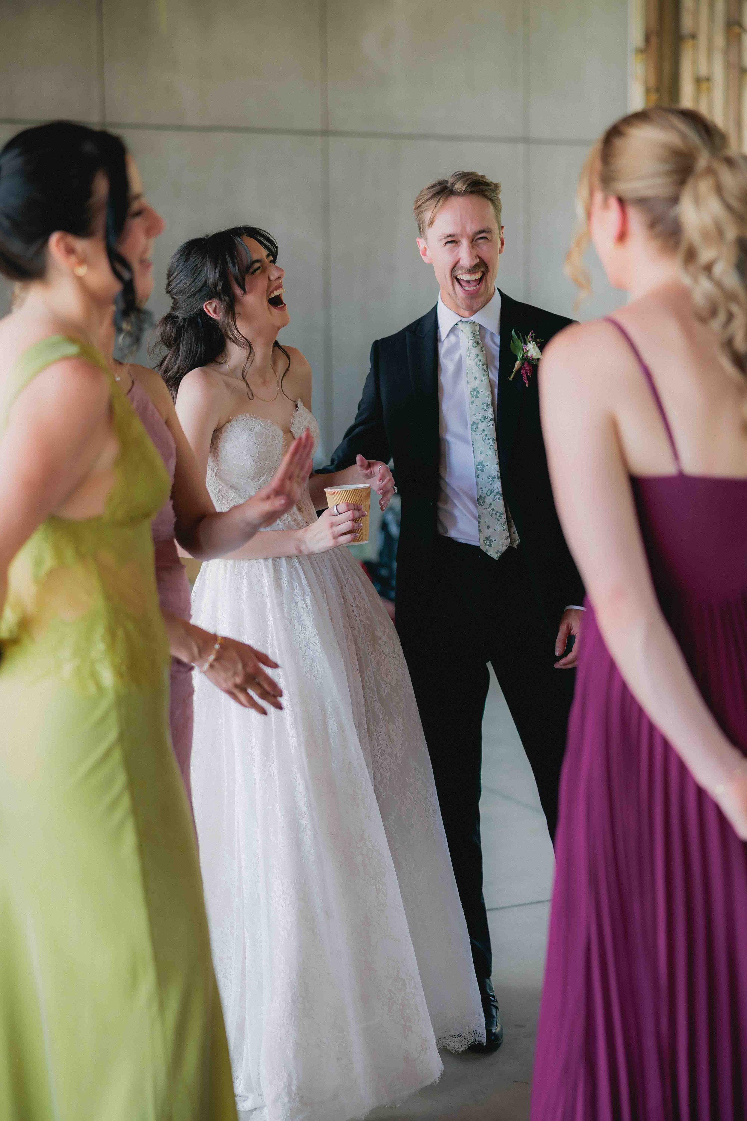 A group of people at a wedding, including a bride in a white wedding dress, a groom in a dark suit, and several women in colorful dresses, standing and laughing together.