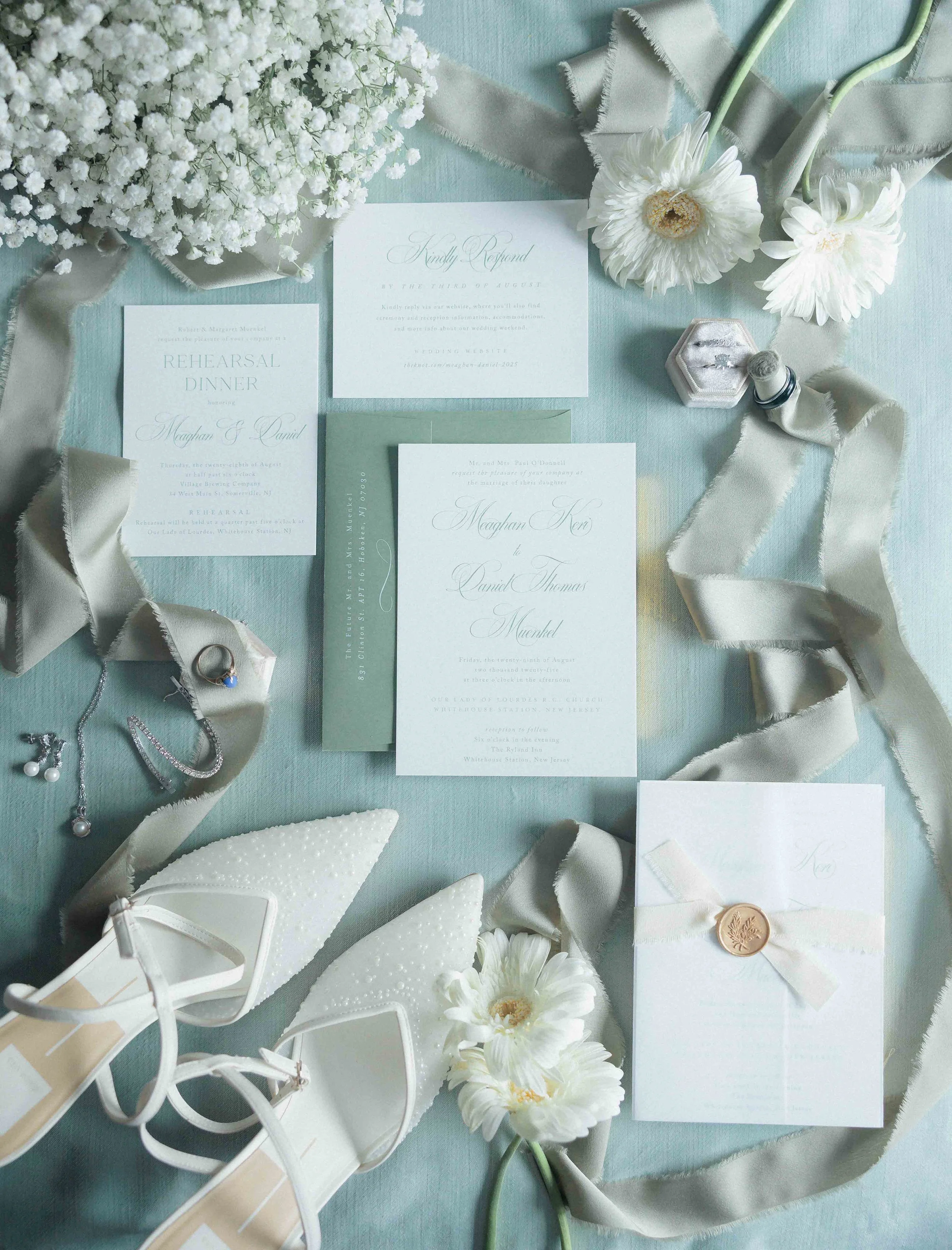Wedding invitation suite with white flowers, pearl jewelry, white high heels, and a satin ribbon on a light blue surface.