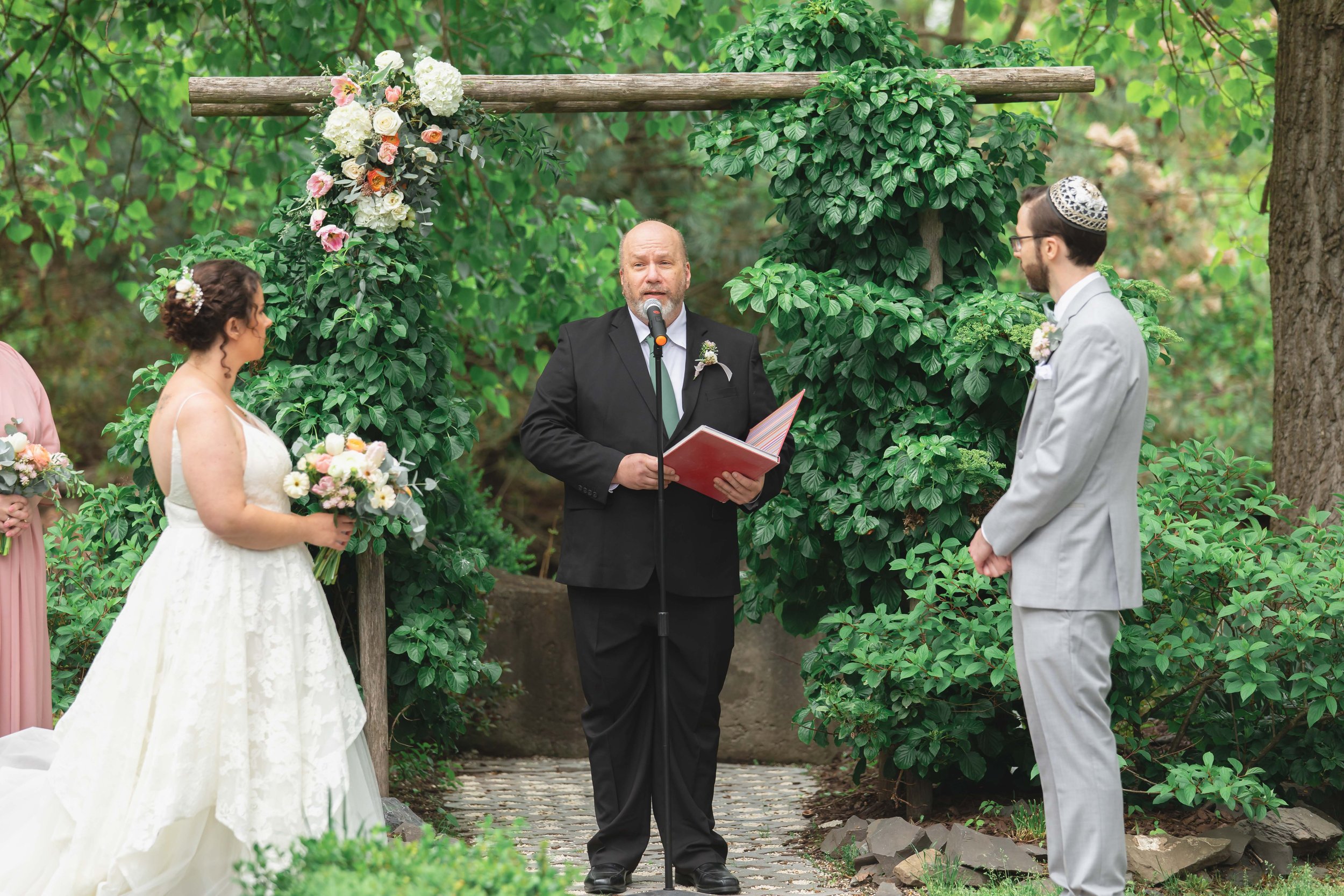 bride and groom under floral decorated arbor in verdant summer garden at Saugerties Performing Arts Factory chic industrial wedding venue in upstate NY