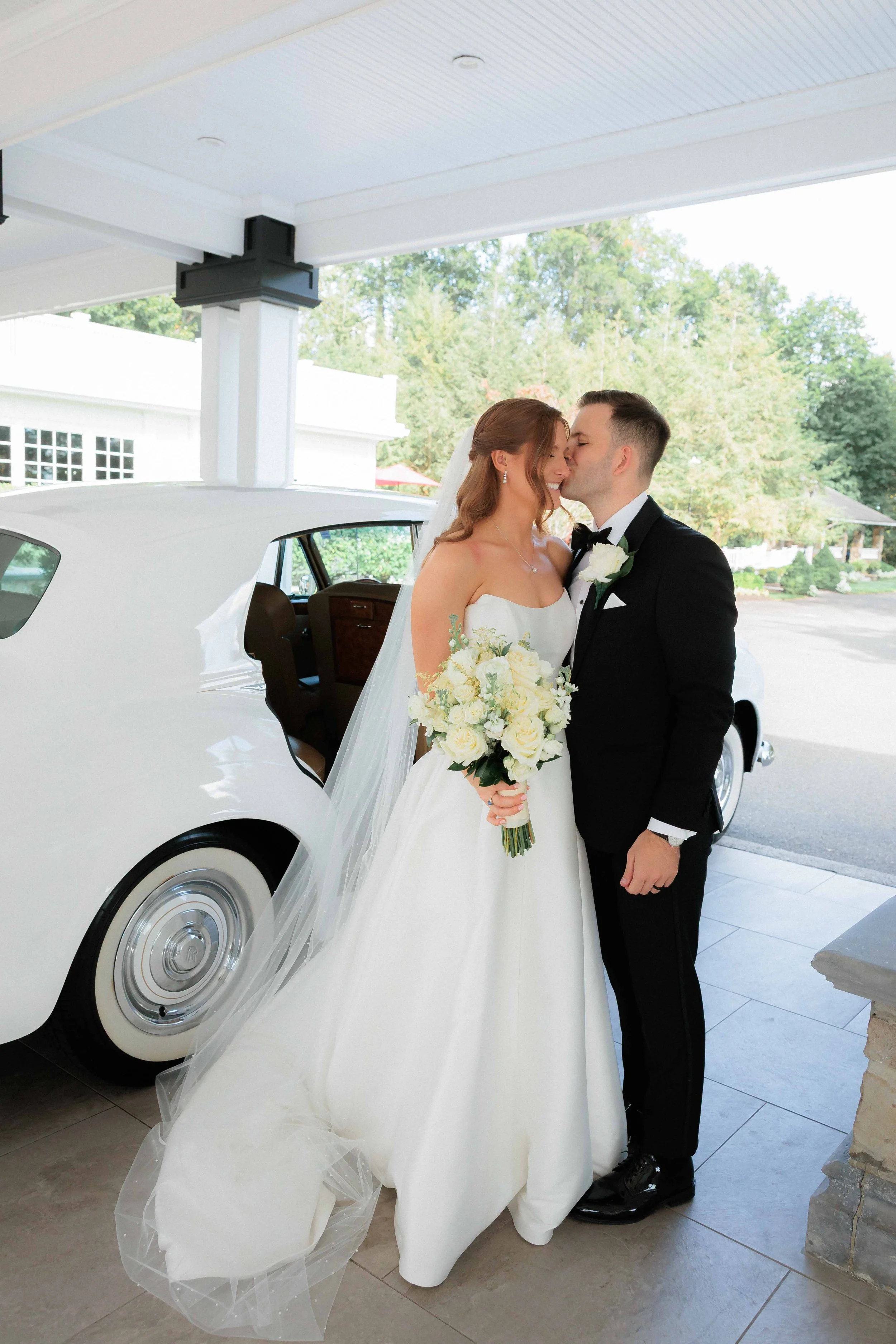 Bride and groom kissing under a carport after their wedding, with the bride holding a bouquet of white roses and dressed in a white gown, and the groom in a black tuxedo.
