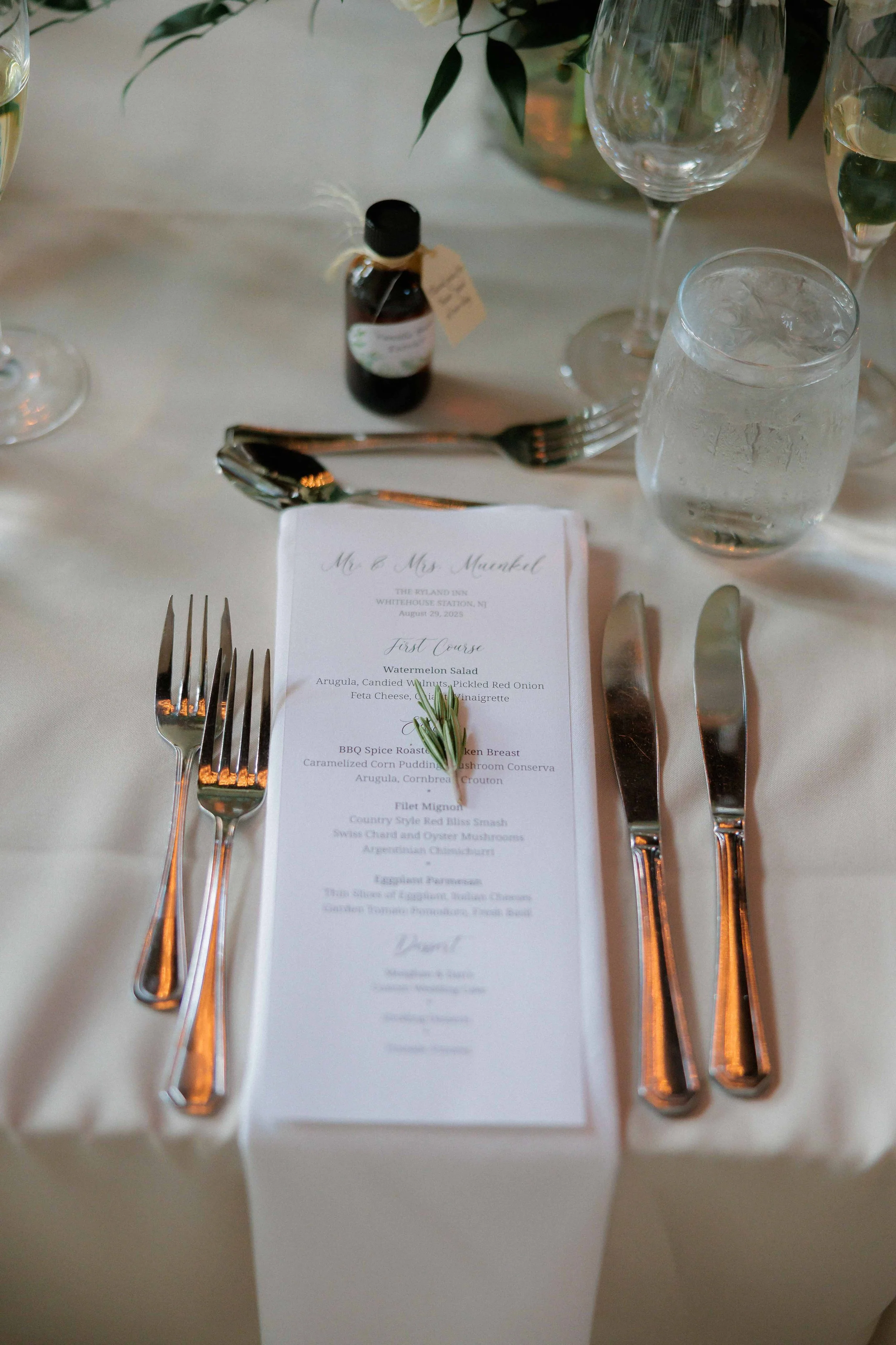Wedding table setting with a printed menu, three forks on the left, three knives on the right, a sprig of rosemary on the menu, a water glass, a wine glass, a glass of water, a small bottle of medicine or syrup, and a floral centerpiece in the backgr