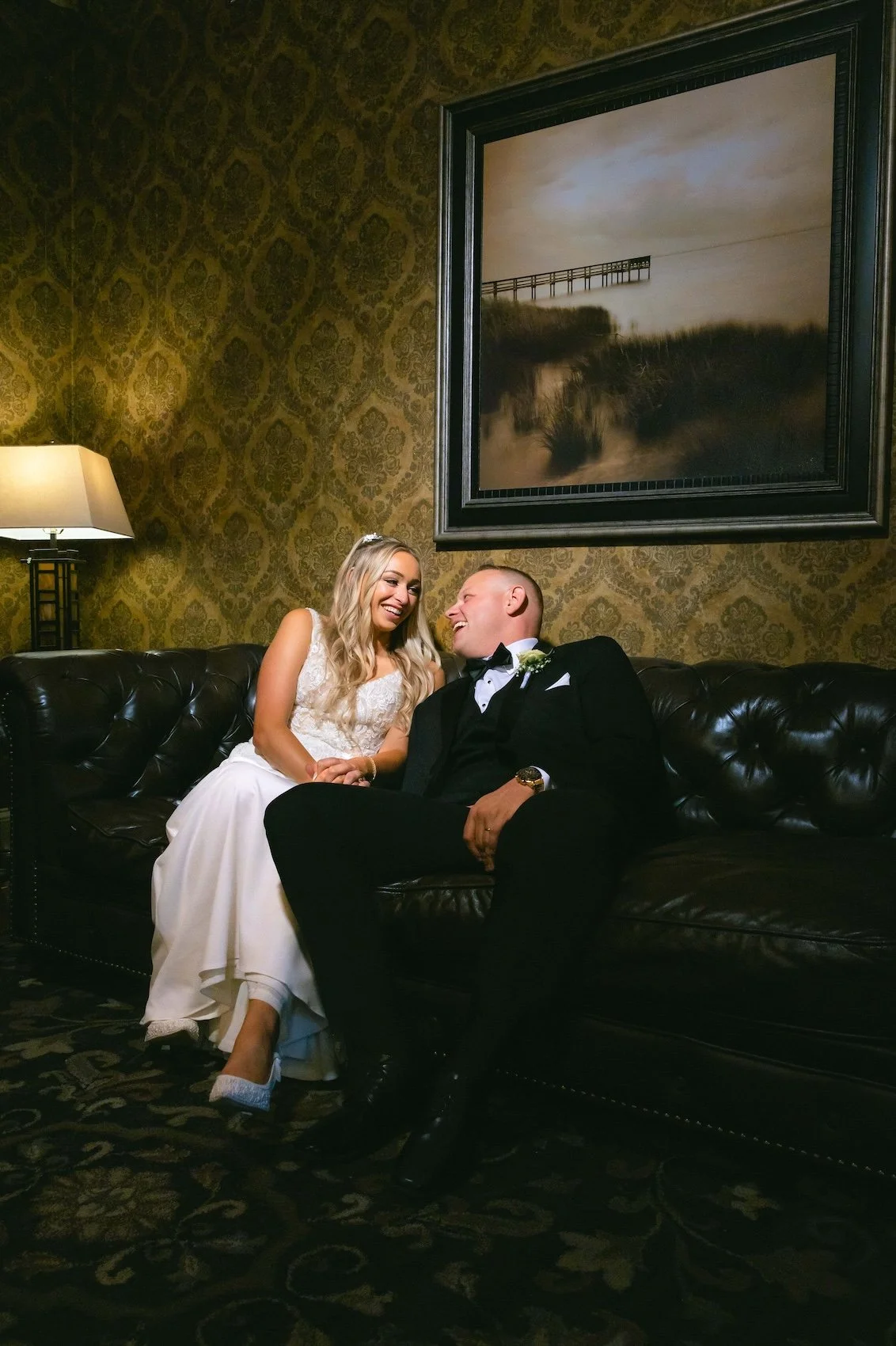 A bride and groom sitting on a black leather couch inside The Old Daley on Crooked Lake wedding venue, smiling and looking at each other, with a framed painting of a pier over water hanging on a patterned wallpapered wall behind them.