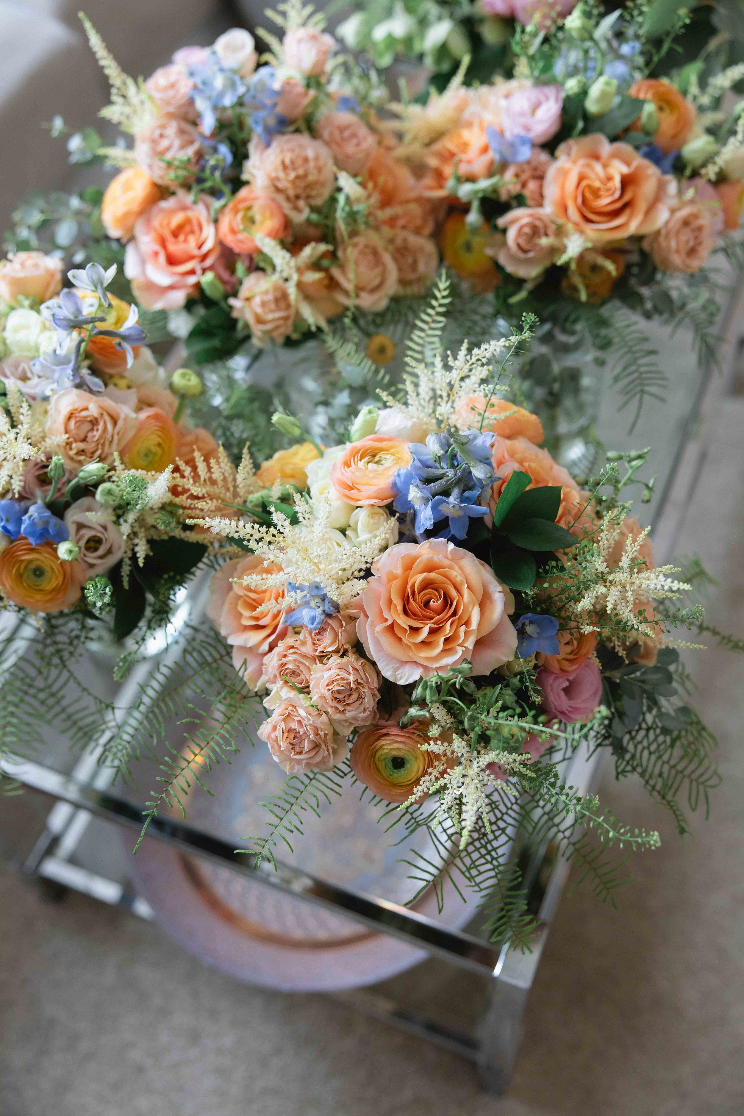 Arrangement of peach, pink, and blue flowers with green fern leaves on a glass table.