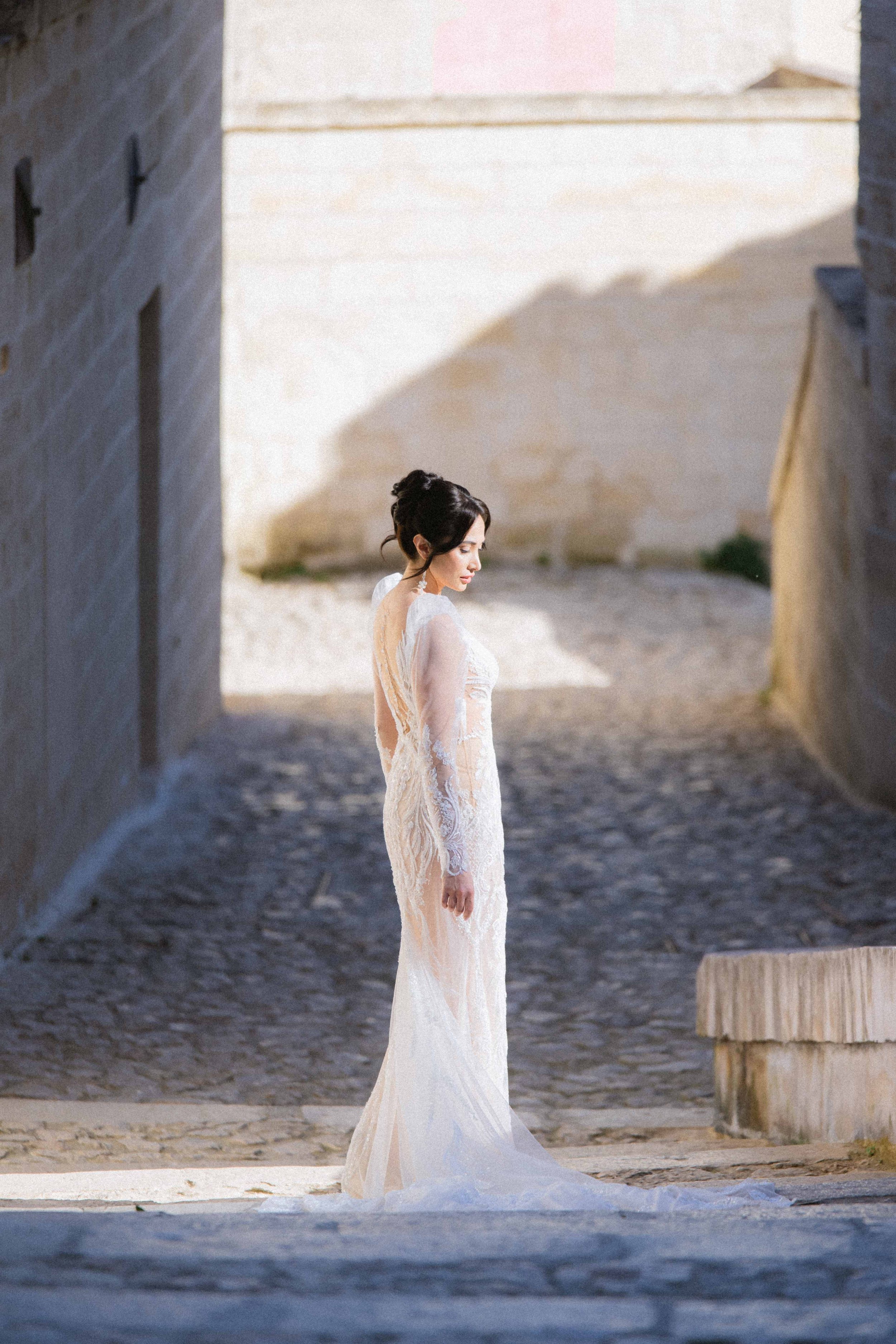 A woman in a white lace wedding dress standing on a stone path between stone walls, looking down with a contemplative expression, illuminated by sunlight.