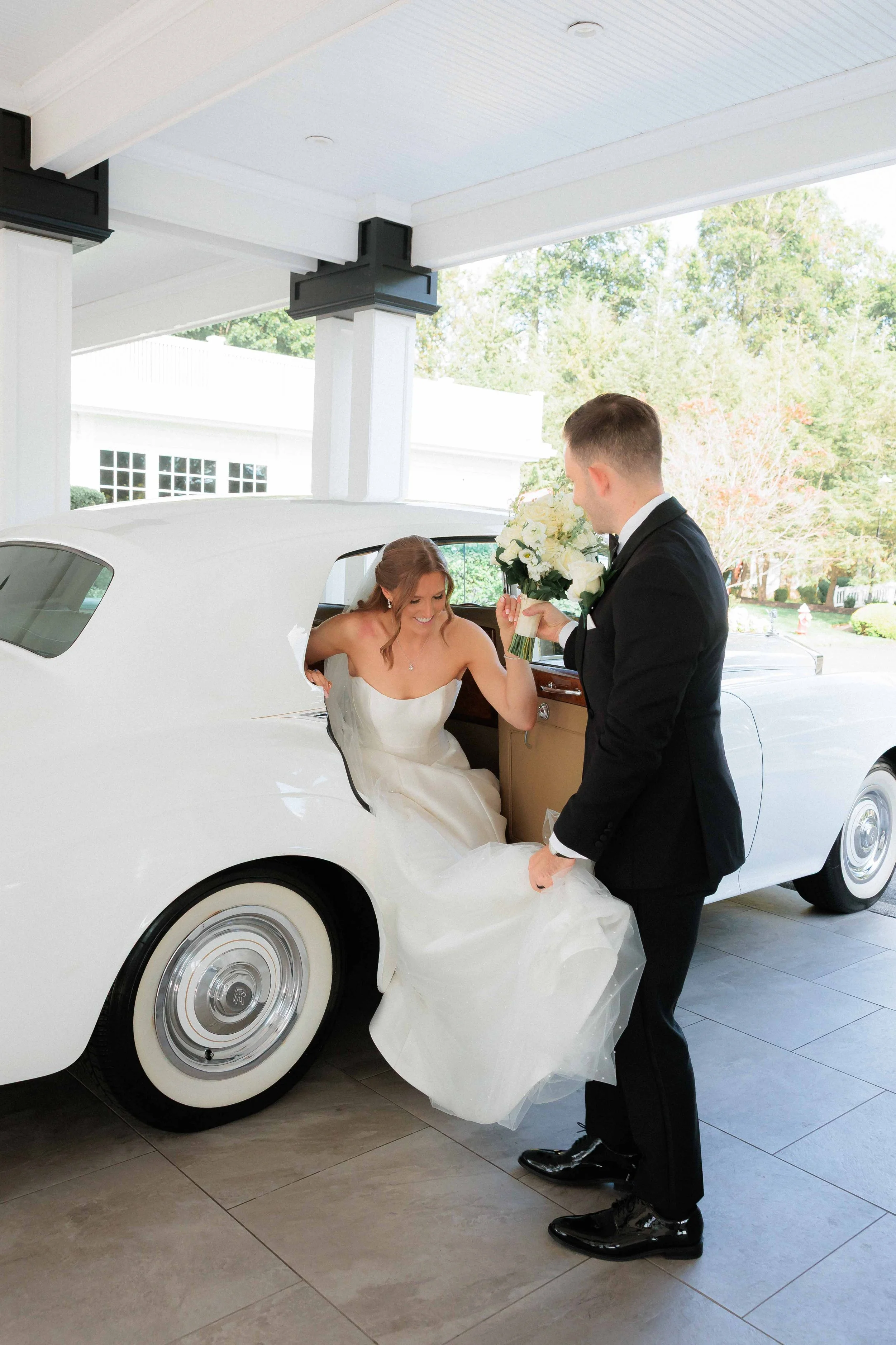 A bride exiting a vintage white car while a groom offers her a bouquet of white flowers at a wedding.