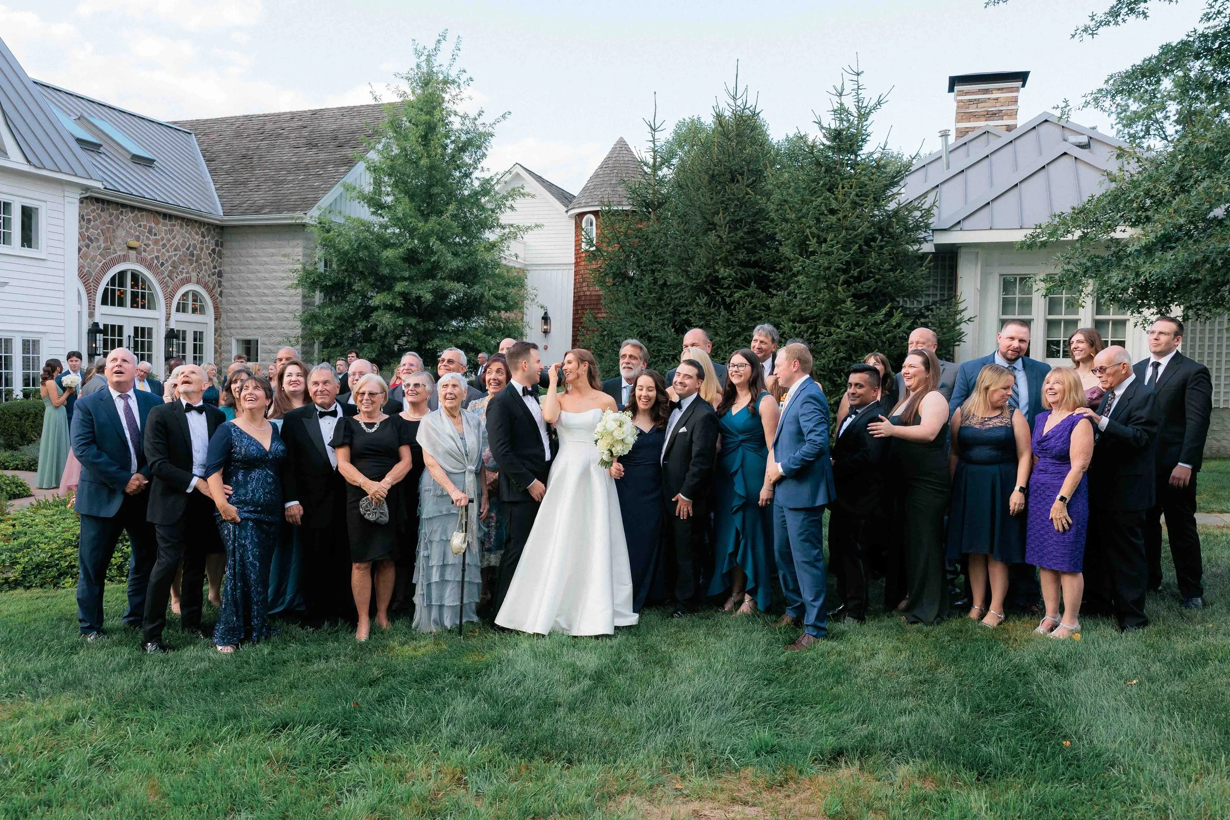 A large group of wedding guests gathered on a lawn, with a bride in a white gown and a groom in a tuxedo in the center, surrounded by family and friends, outdoors in front of a house with trees and a cloudy sky.