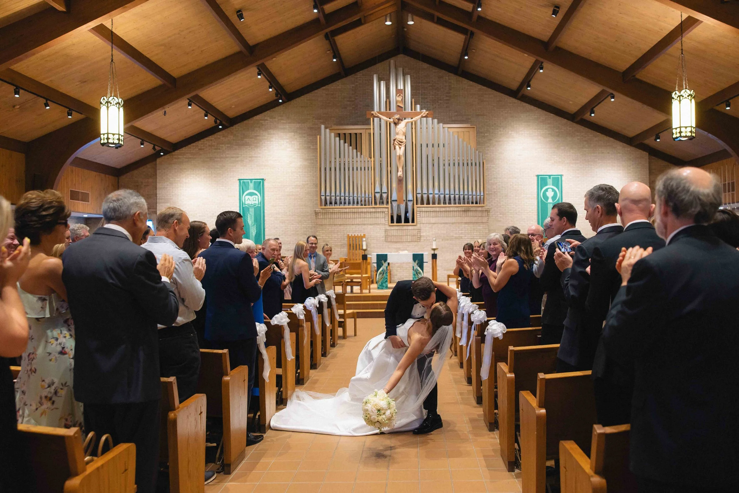 A bride and groom kiss at their wedding altar in a church with pews decorated with white ribbons, while guests clap and take photos. The church features a wooden ceiling, hanging lights, a large crucifix, and organ pipes behind the altar.