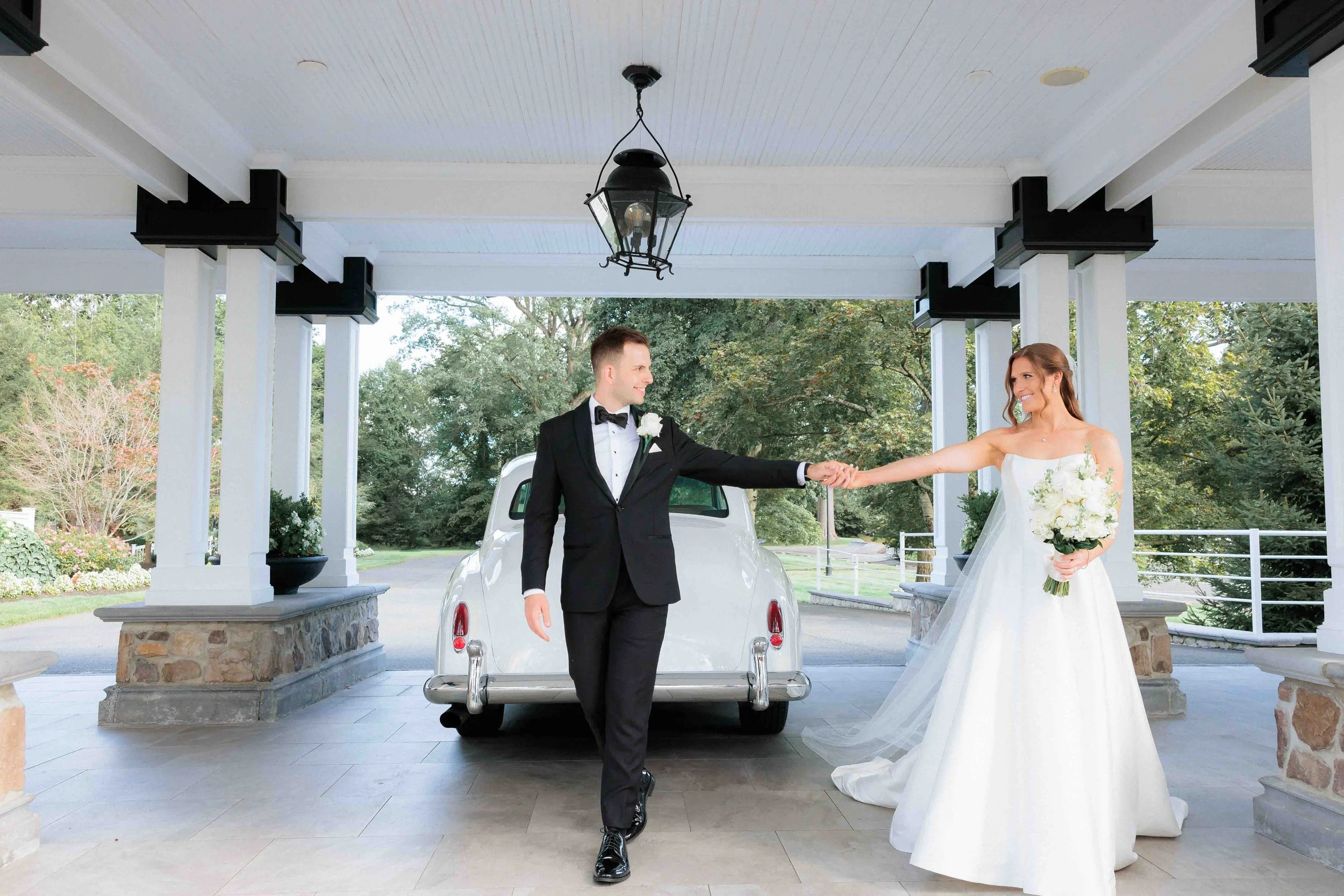 A newlywed couple standing on a porch, with a white vintage car behind them. The groom in a black tuxedo with a bow tie is reaching out to hold the bride's hand, who is in a white wedding gown holding a bouquet of white flowers.
