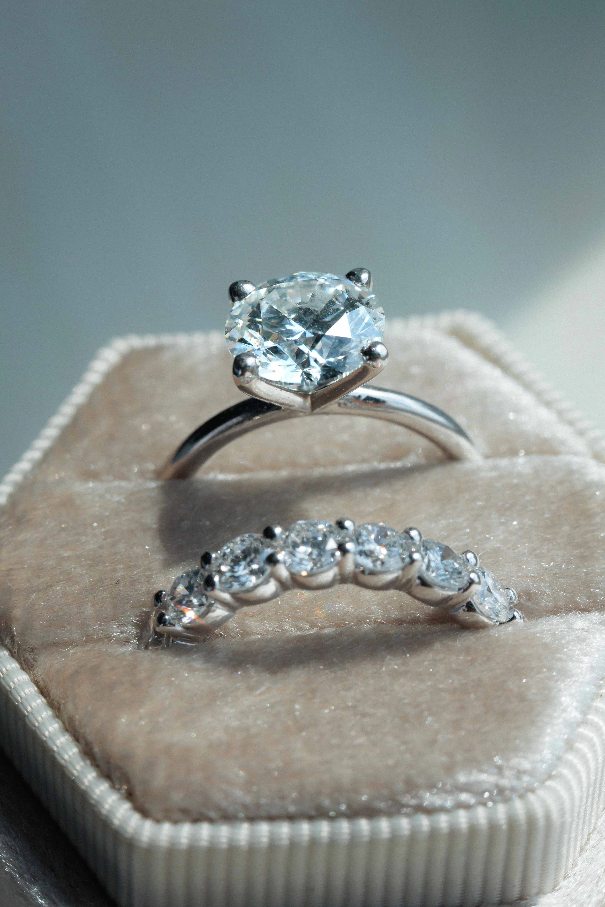 A close-up of a solitaire diamond engagement ring and a diamond eternity band resting on a soft beige ring box.