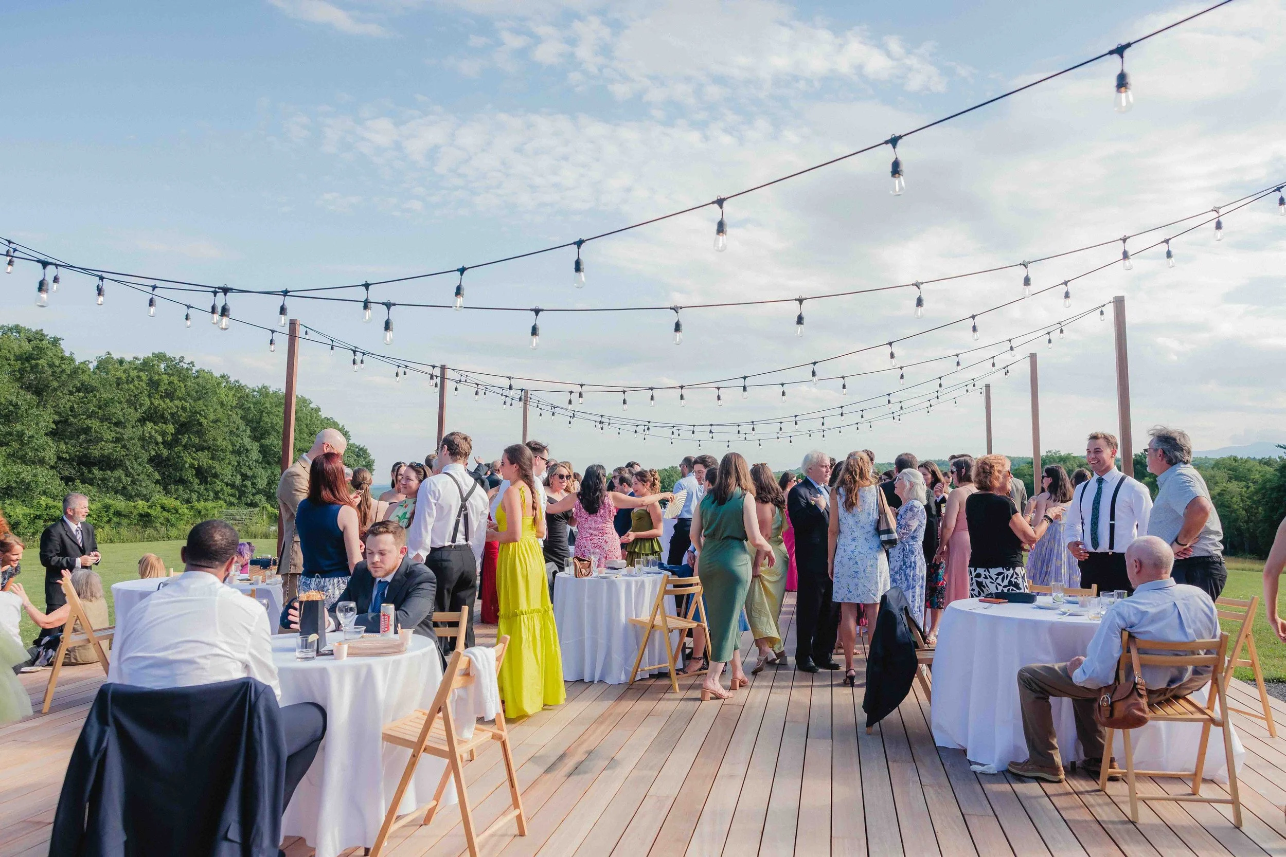 Outdoor wedding reception with guests mingling, some seated at tables with white tablecloths, and others standing and chatting. String lights hang above, and lush greenery is visible in the background.