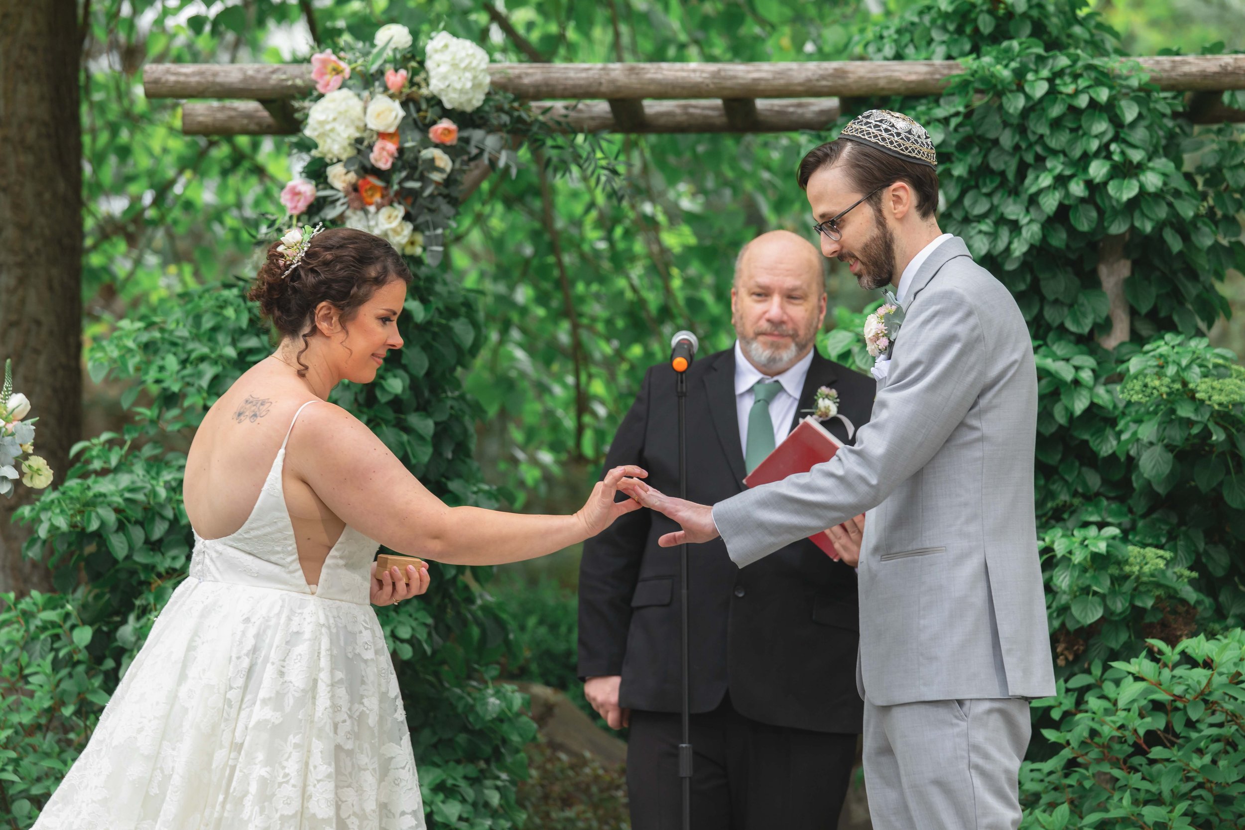 bride puts groom's ring on finger under floral decorated arbor in verdant summer garden at Saugerties Performing Arts Factory chic industrial wedding venue in upstate NY
