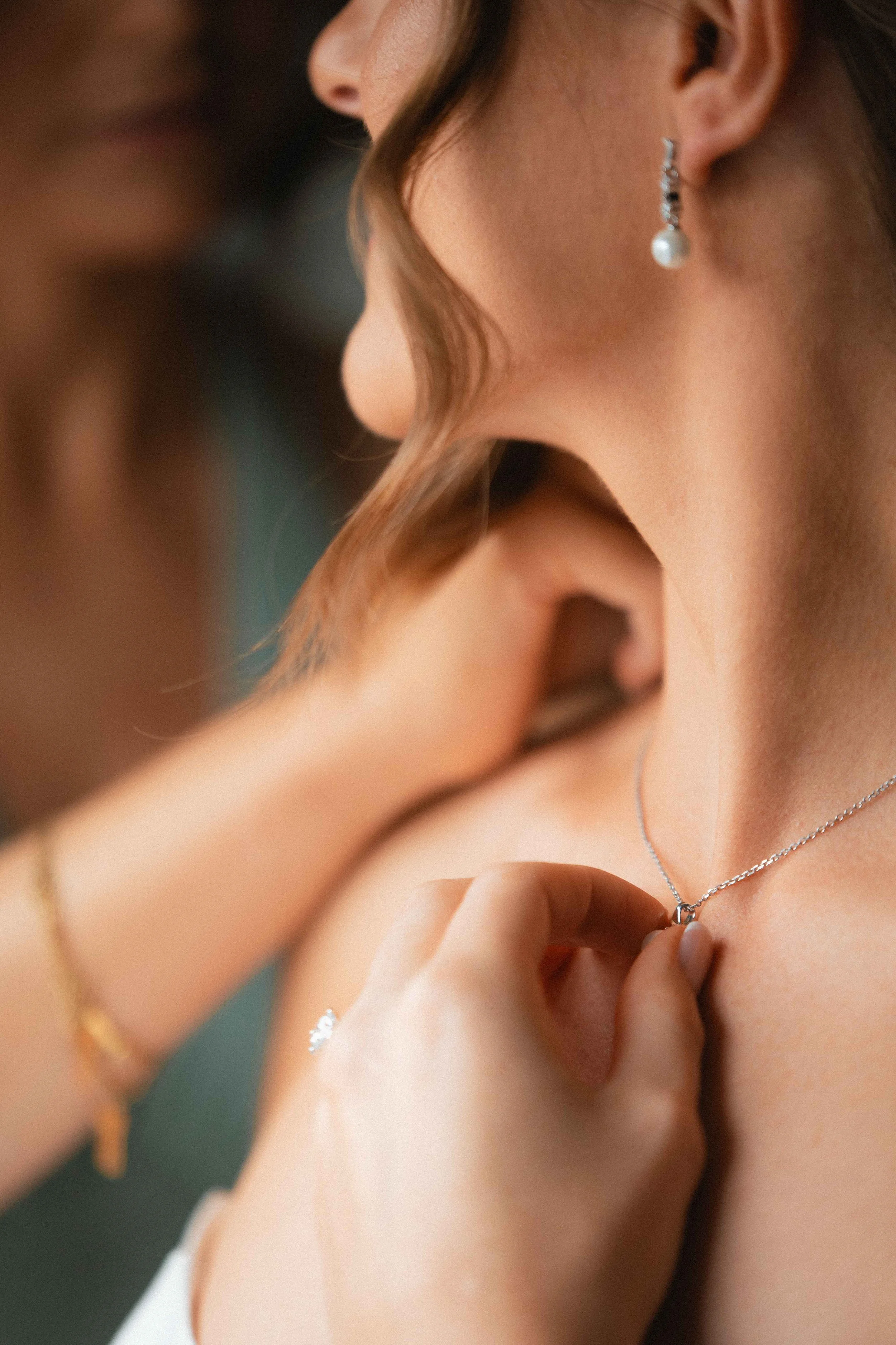 Close-up of a woman wearing pearl earrings and a silver necklace, with a person adjusting the jewelry on her neck.
