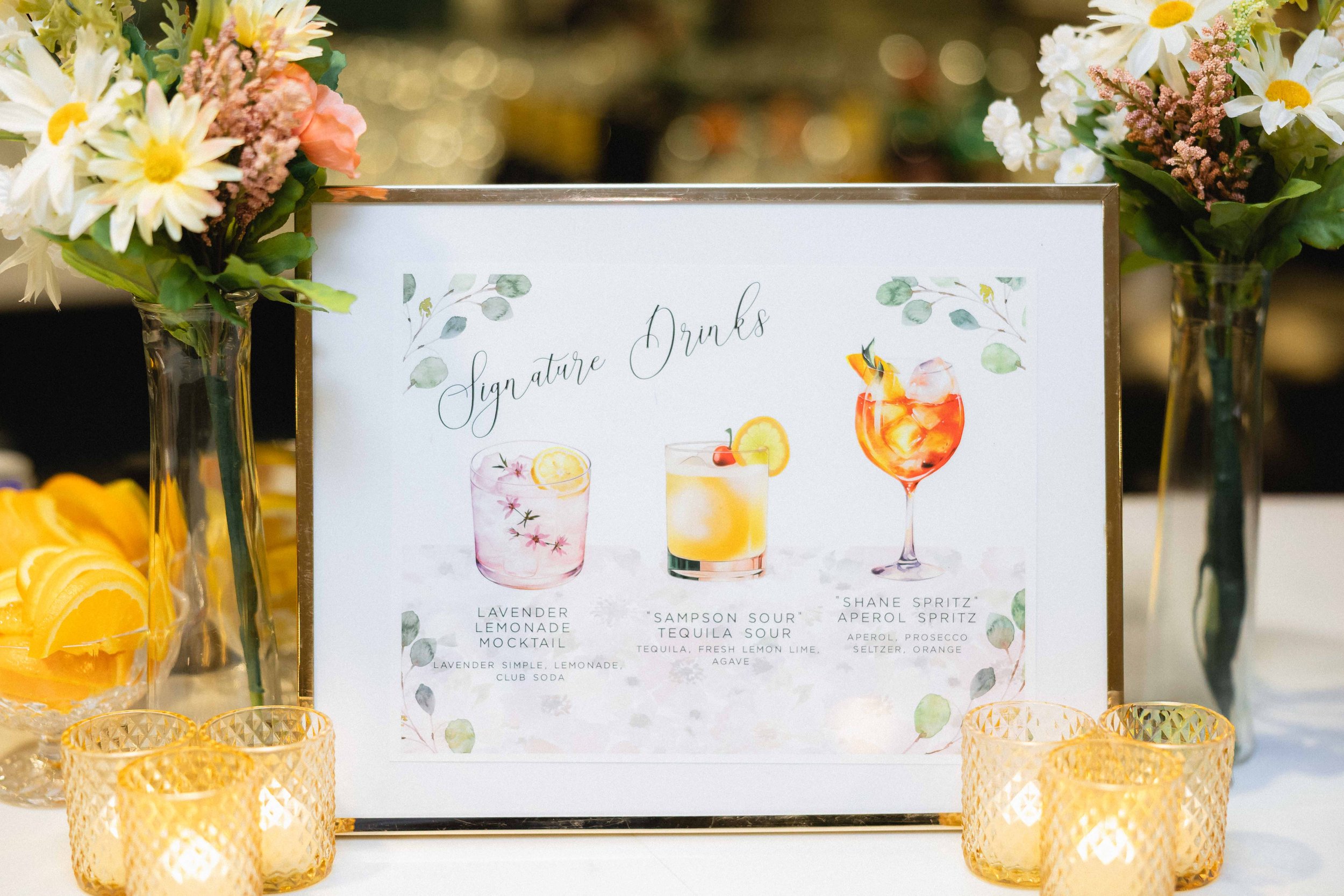 A framed menu sign for signature drinks, featuring illustrations of three cocktails: Lavender Lemonade Mocktail, Sampson Sour Tequila Sour, and Shane Spritz Aperol Spritz. The sign is decorated with watercolor green leaves and flanked by two vases of