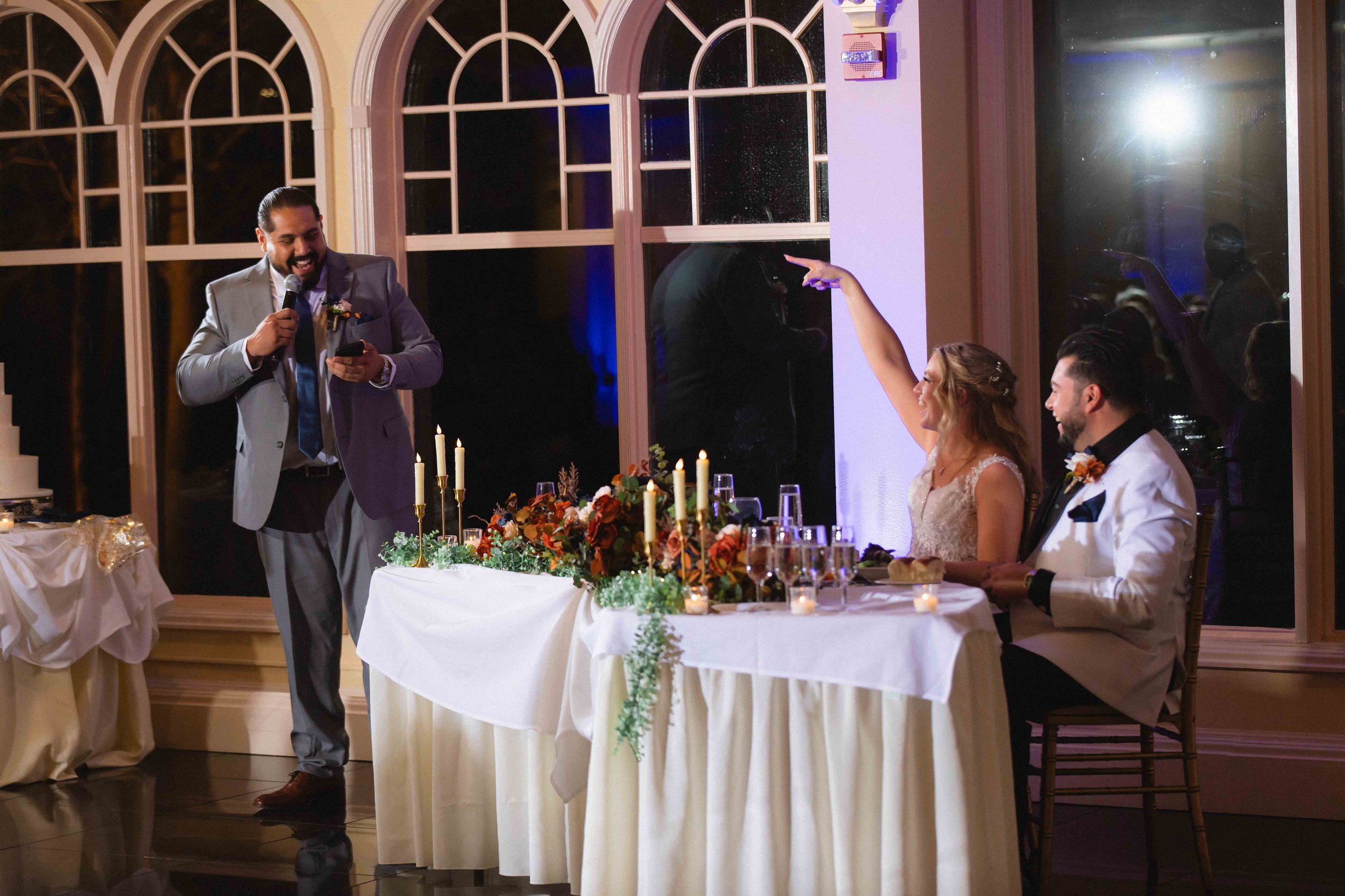 bride and groom laughing and pointing at groom's brother who is giving toast speech in front of newlywed's table during reception at the waterview wedding venue in monroe ct