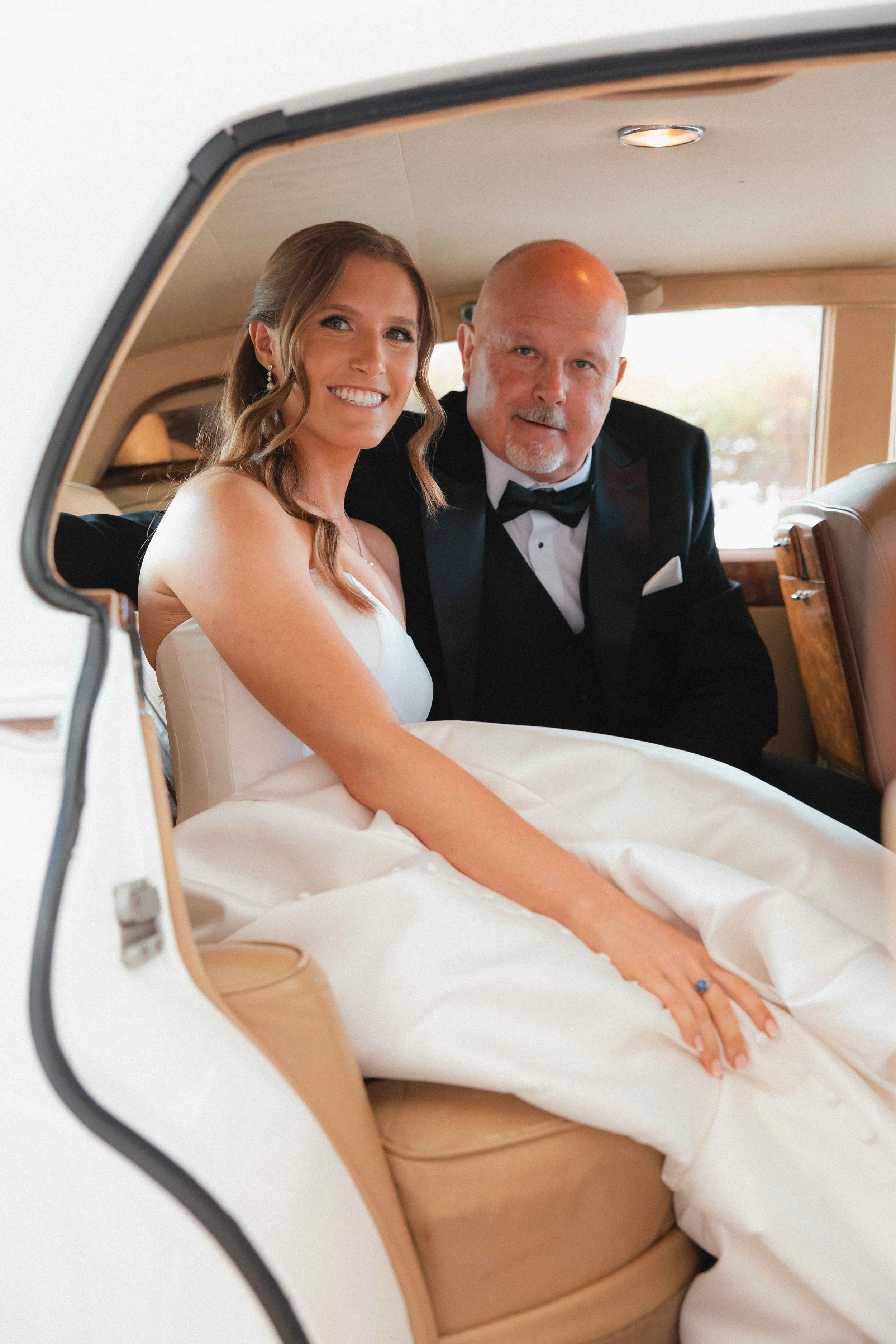 A bride sitting inside a vintage car with an older man, both dressed formally for a wedding.
