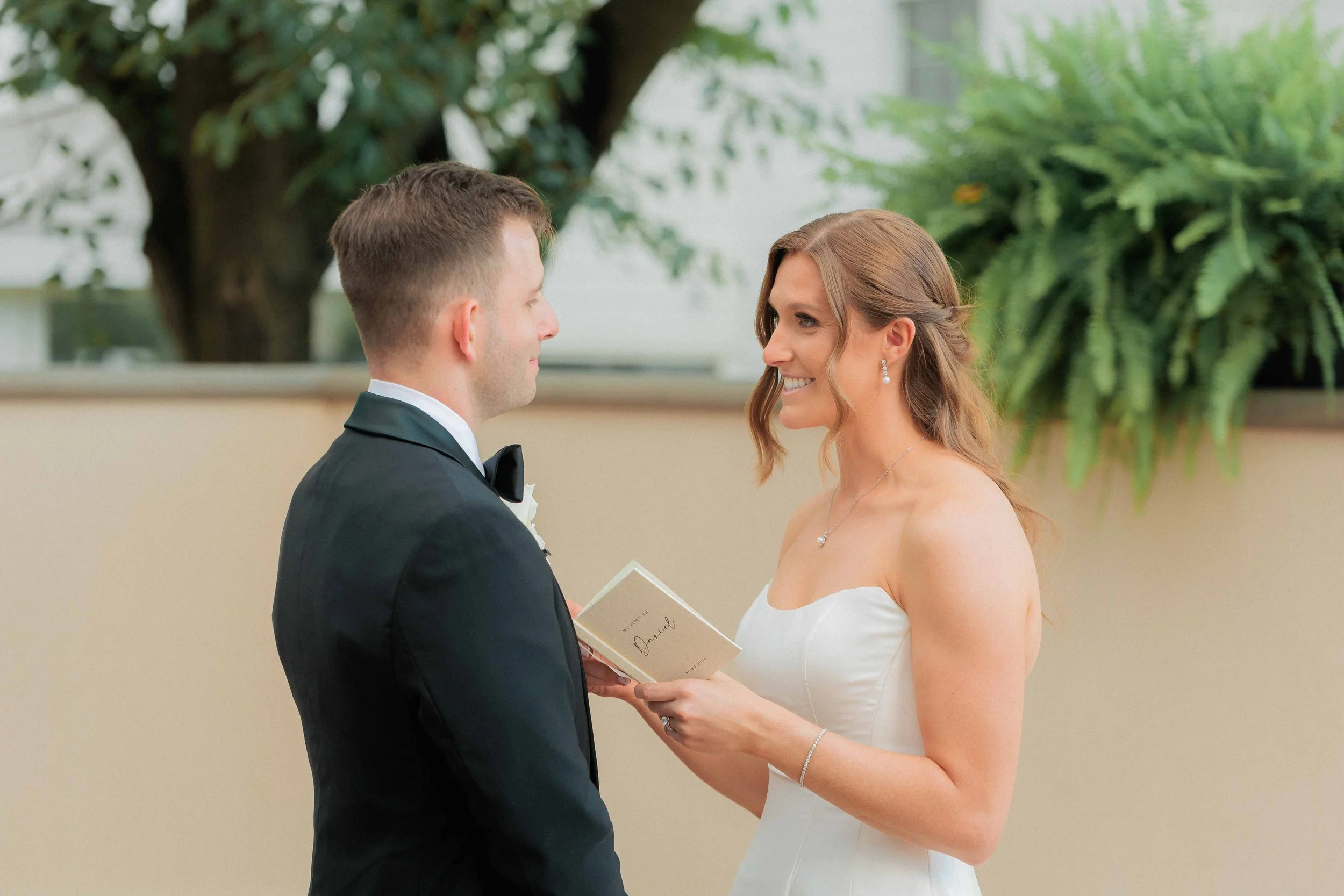 A bride and groom exchanging vows in a wedding ceremony indoors, with the bride smiling while holding a small booklet.