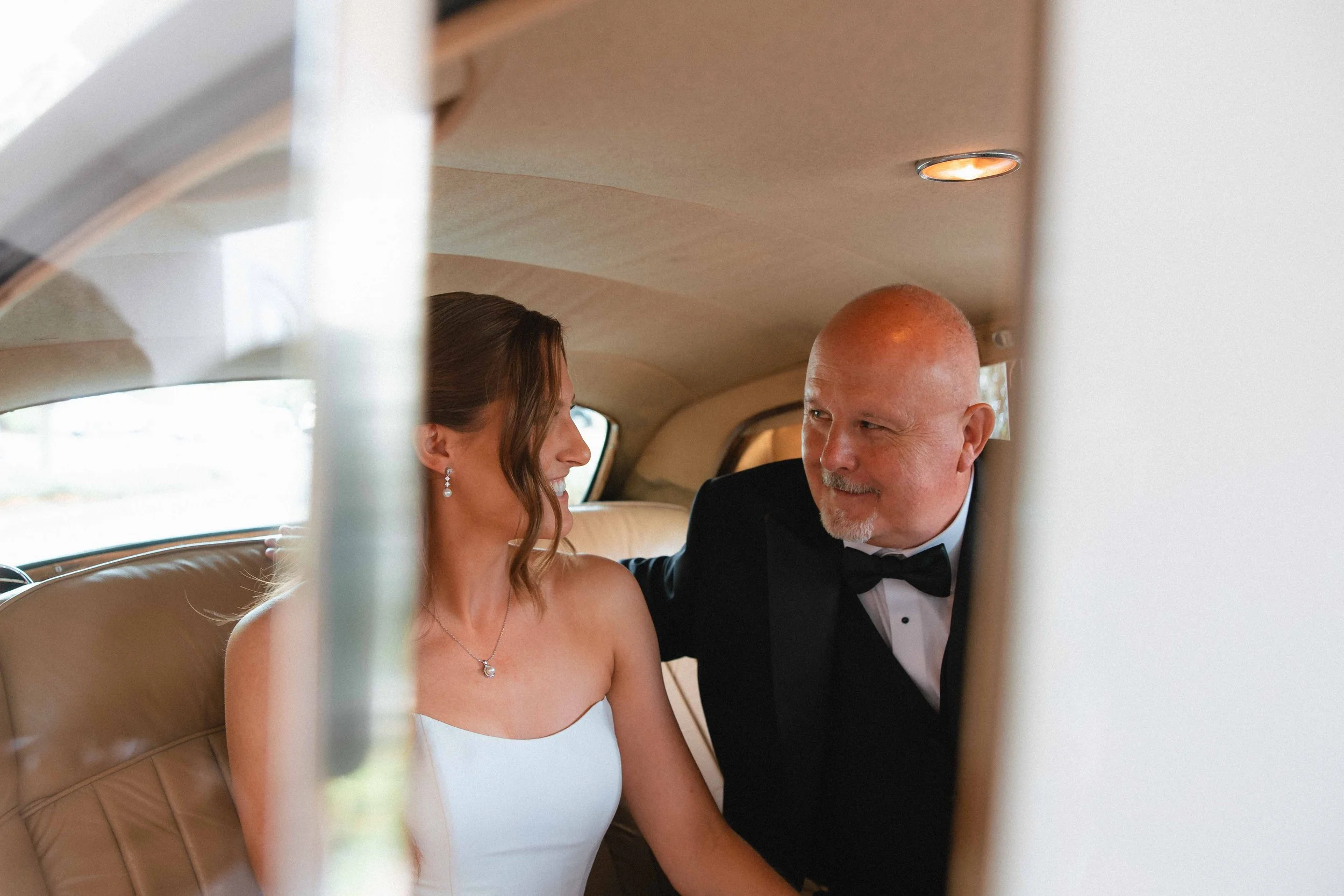 A bride in a white dress and a groom in a tuxedo sitting inside a vintage car, smiling and looking at each other.