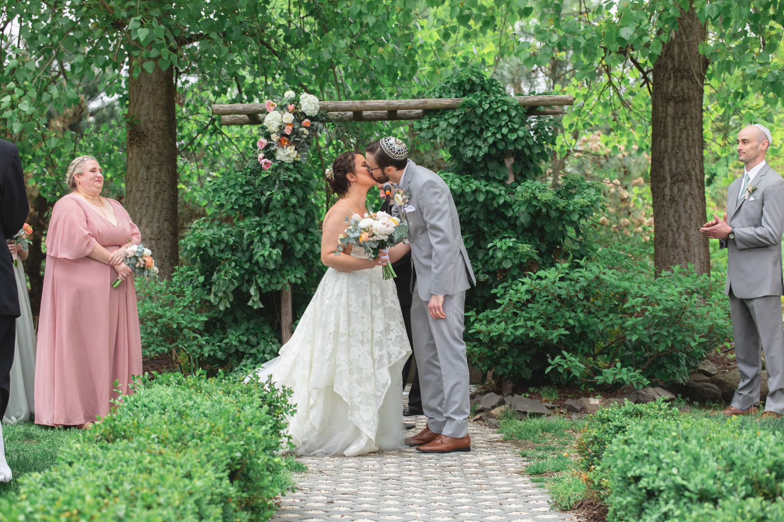 bride and groom kiss under floral decorated arbor in verdant summer garden at Saugerties Performing Arts Factory chic industrial wedding venue in upstate NY