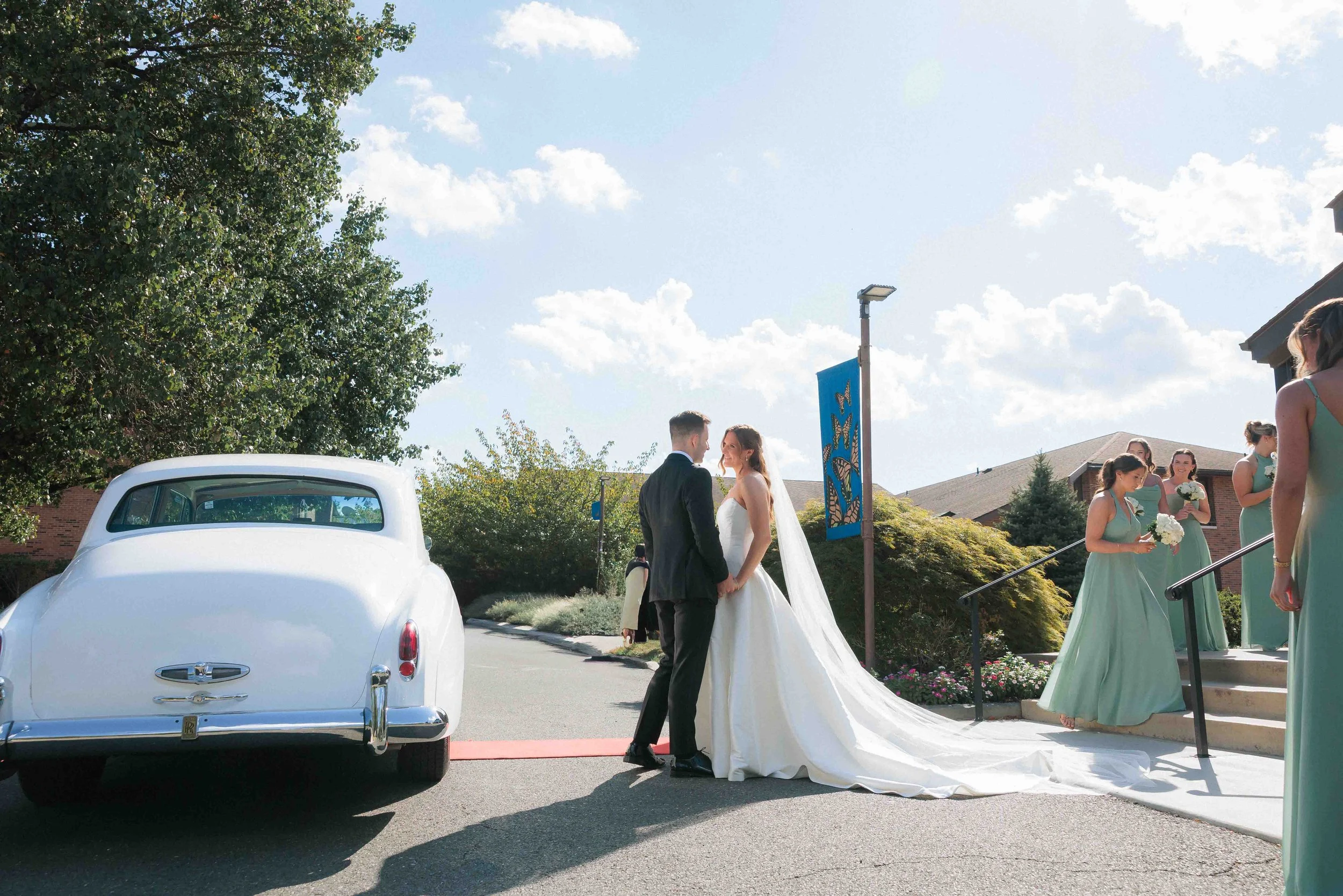 Bride and groom holding hands and smiling in wedding procession, surrounded by bridesmaids holding bouquets, outside on a sunny day.