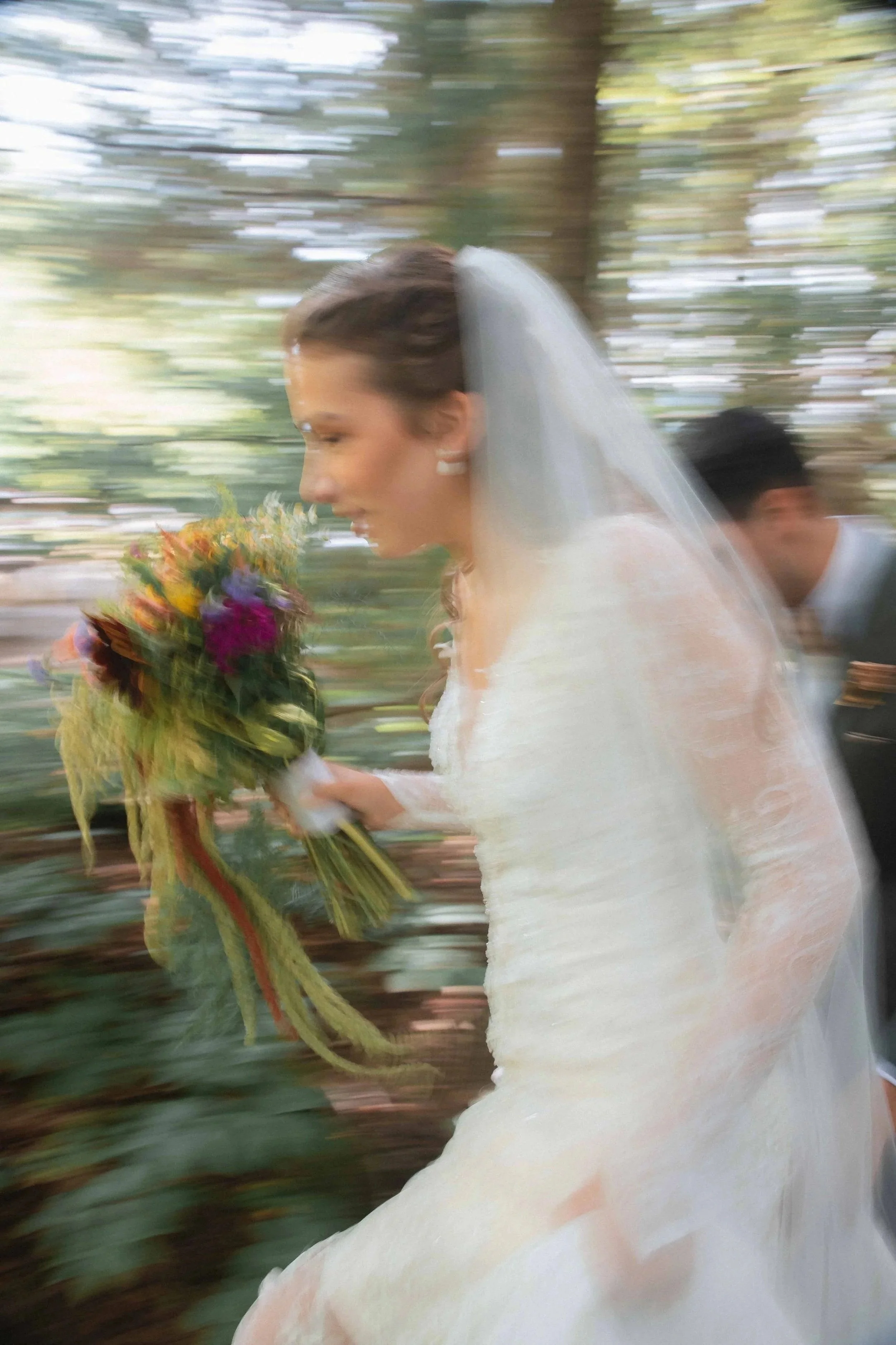 A bride wearing a white dress and veil holding a colorful bouquet of flowers, walking outdoors among trees with a blurred background.