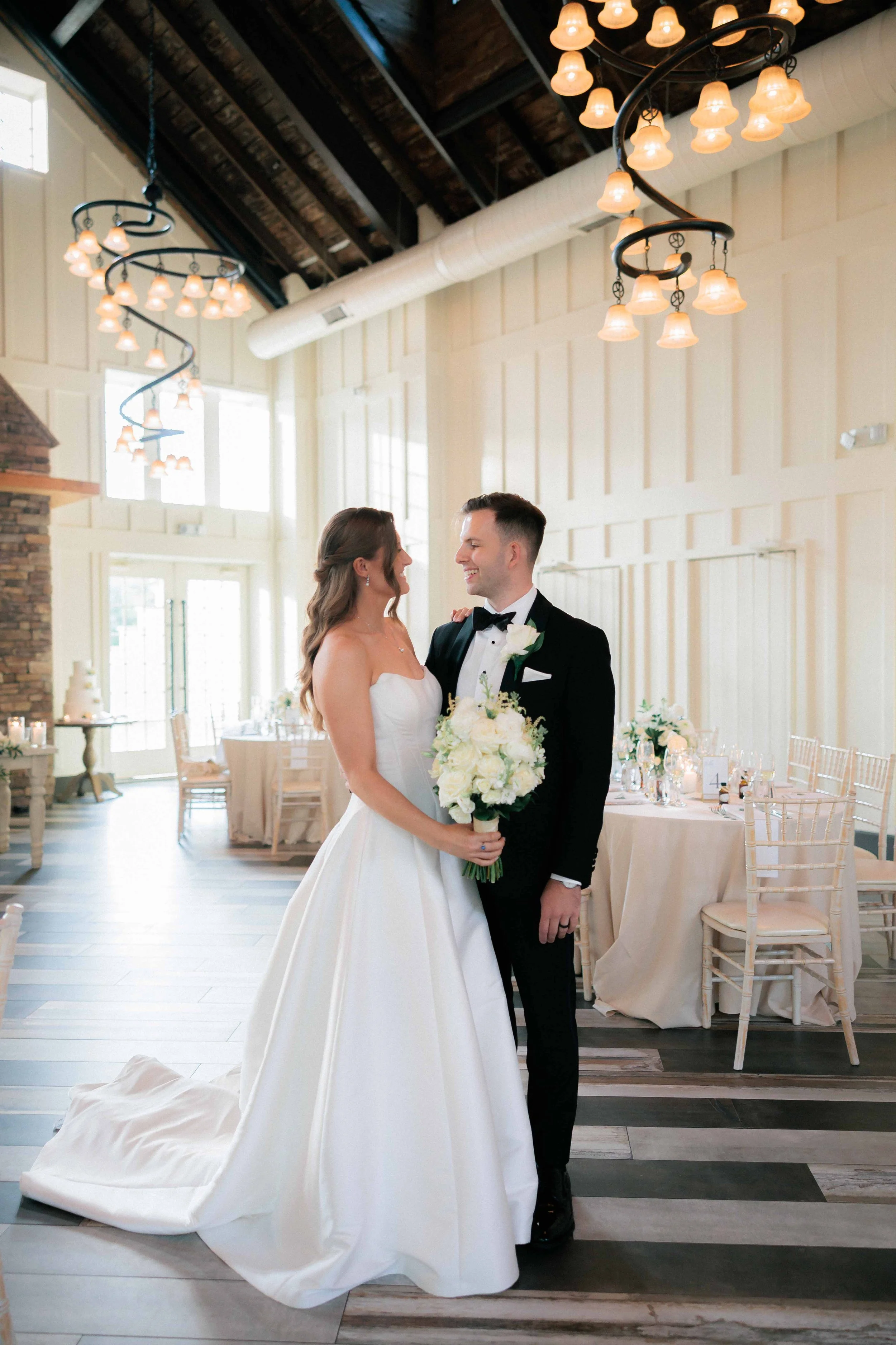 Bride and groom sharing a moment at their wedding reception, with the bride holding a bouquet and wearing a strapless white wedding gown, and the groom dressed in a tuxedo, inside a decorated elegant event space.