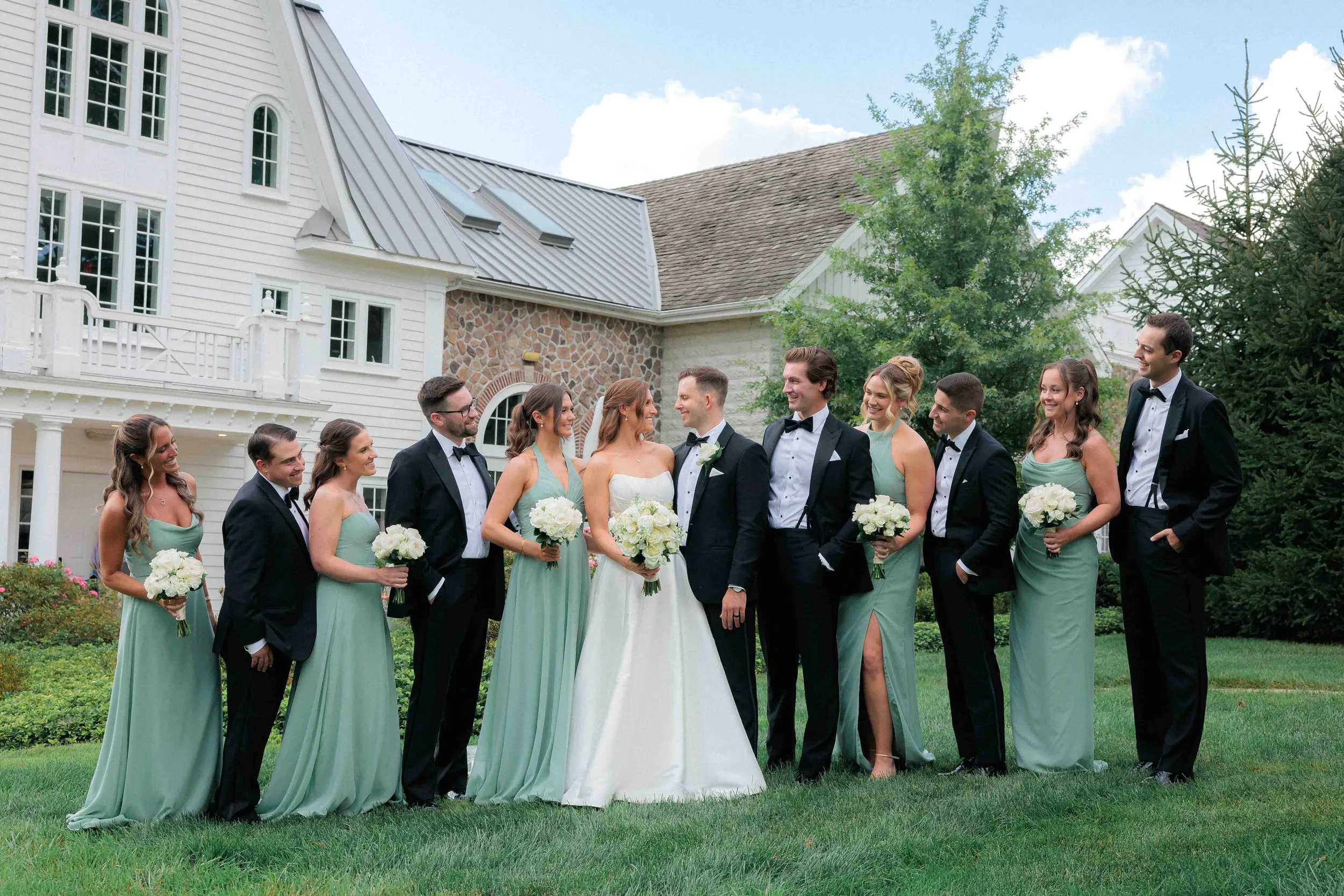A wedding party standing outdoors on a lawn with a large white house in the background. The group consists of a bride and groom in the center, surrounded by bridesmaids in green dresses and groomsmen in black tuxedos. Everyone is smiling and holding