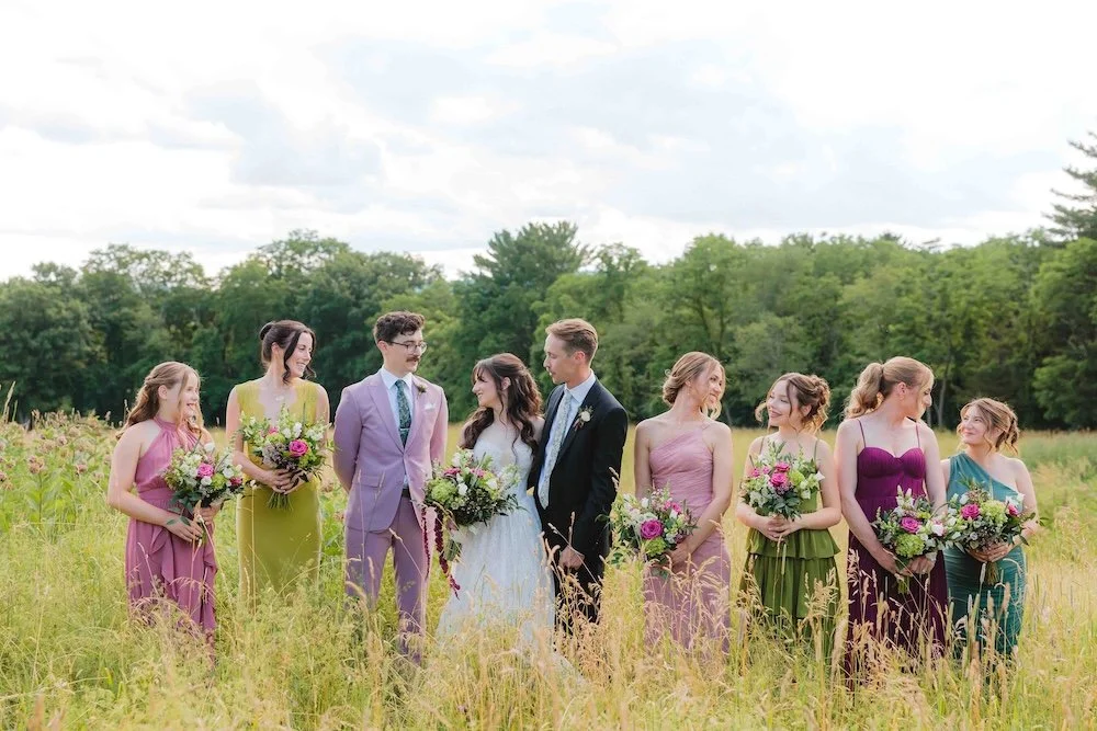 A group of people at a wedding standing in a grassy field with trees in the background. The bride and groom are in the center, surrounded by bridesmaids and groomsmen holding bouquets.