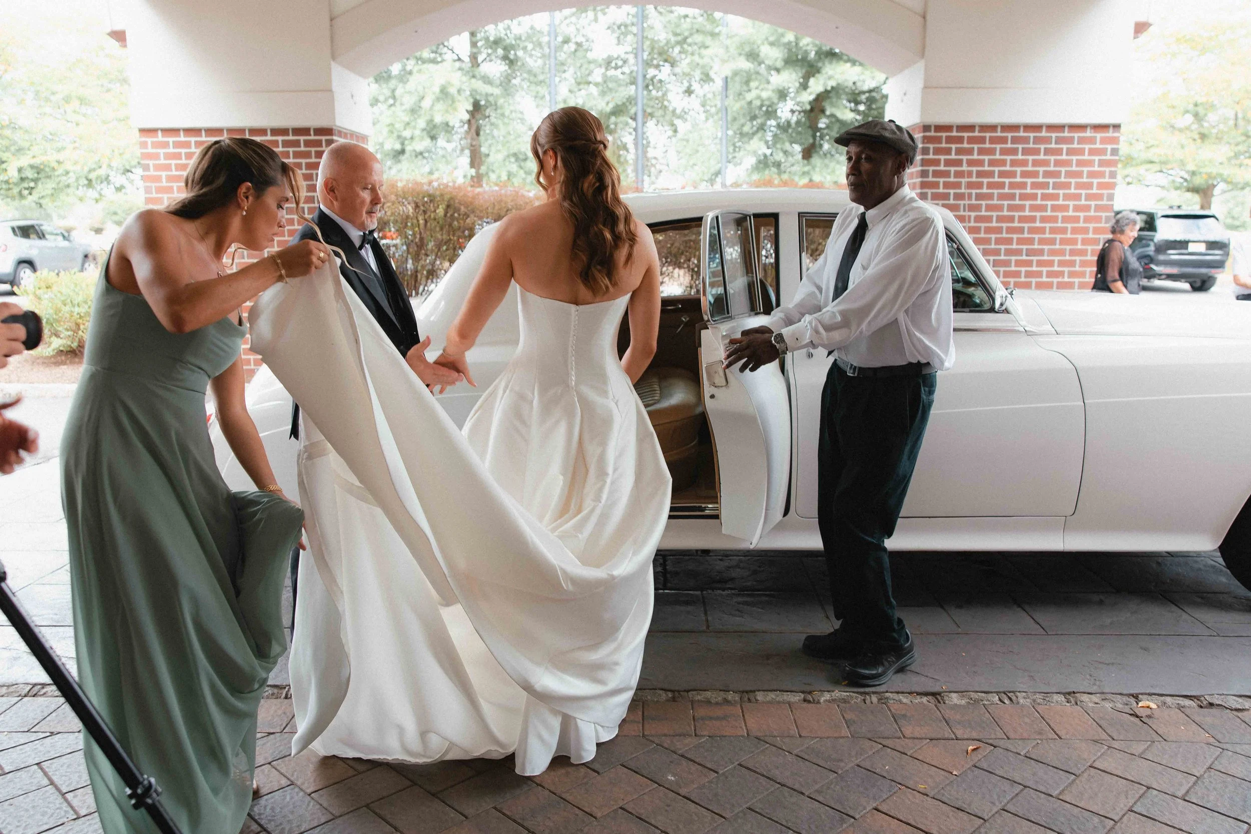 A bride in a strapless wedding dress is stepping out of a white vintage car as attendants help with her gown on a brick-paved area under an archway.