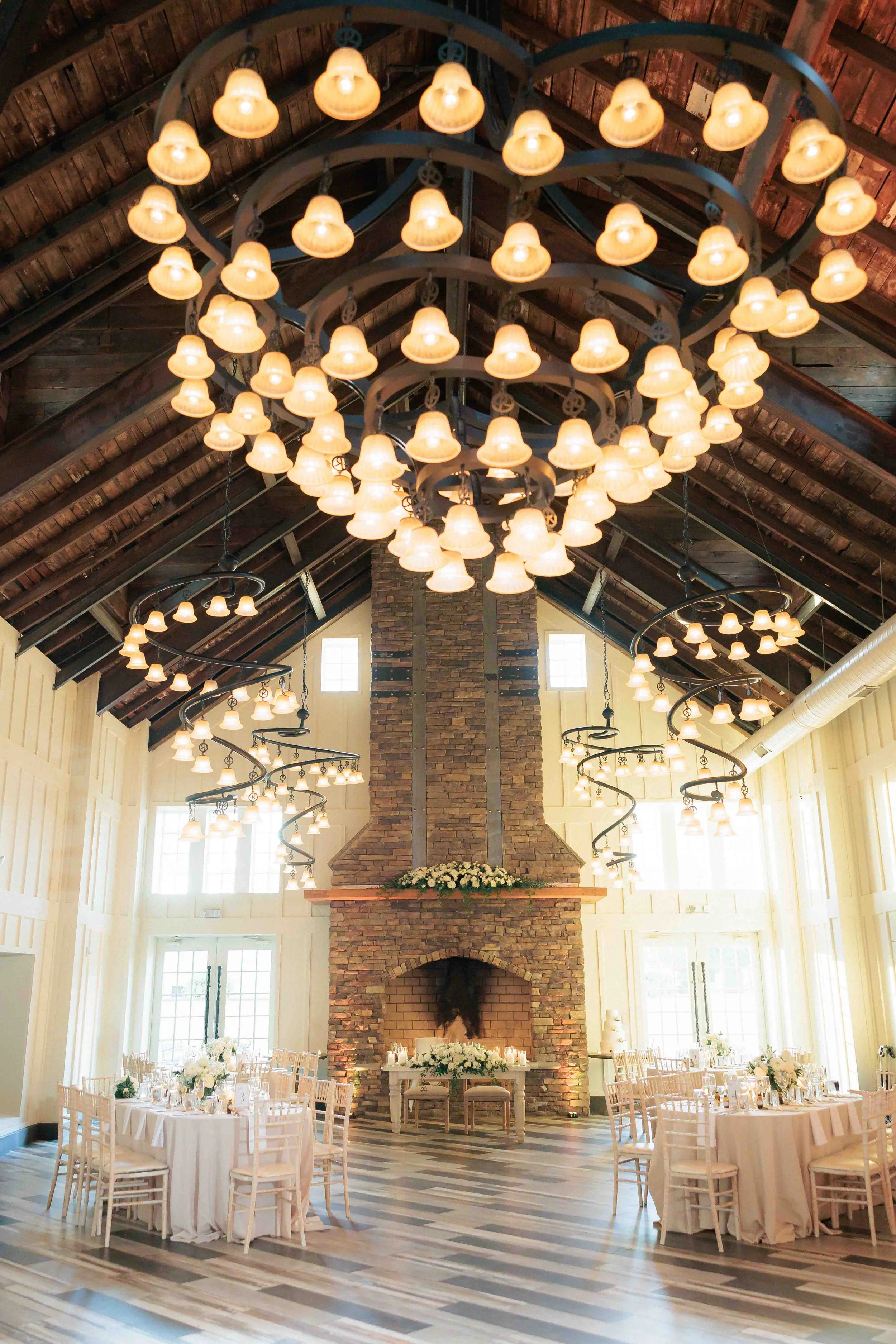 Elegant wedding reception setup in a rustic venue with a large brick fireplace, high vaulted wood ceiling, multiple chandeliers, and round tables with white tablecloths and floral centerpieces.