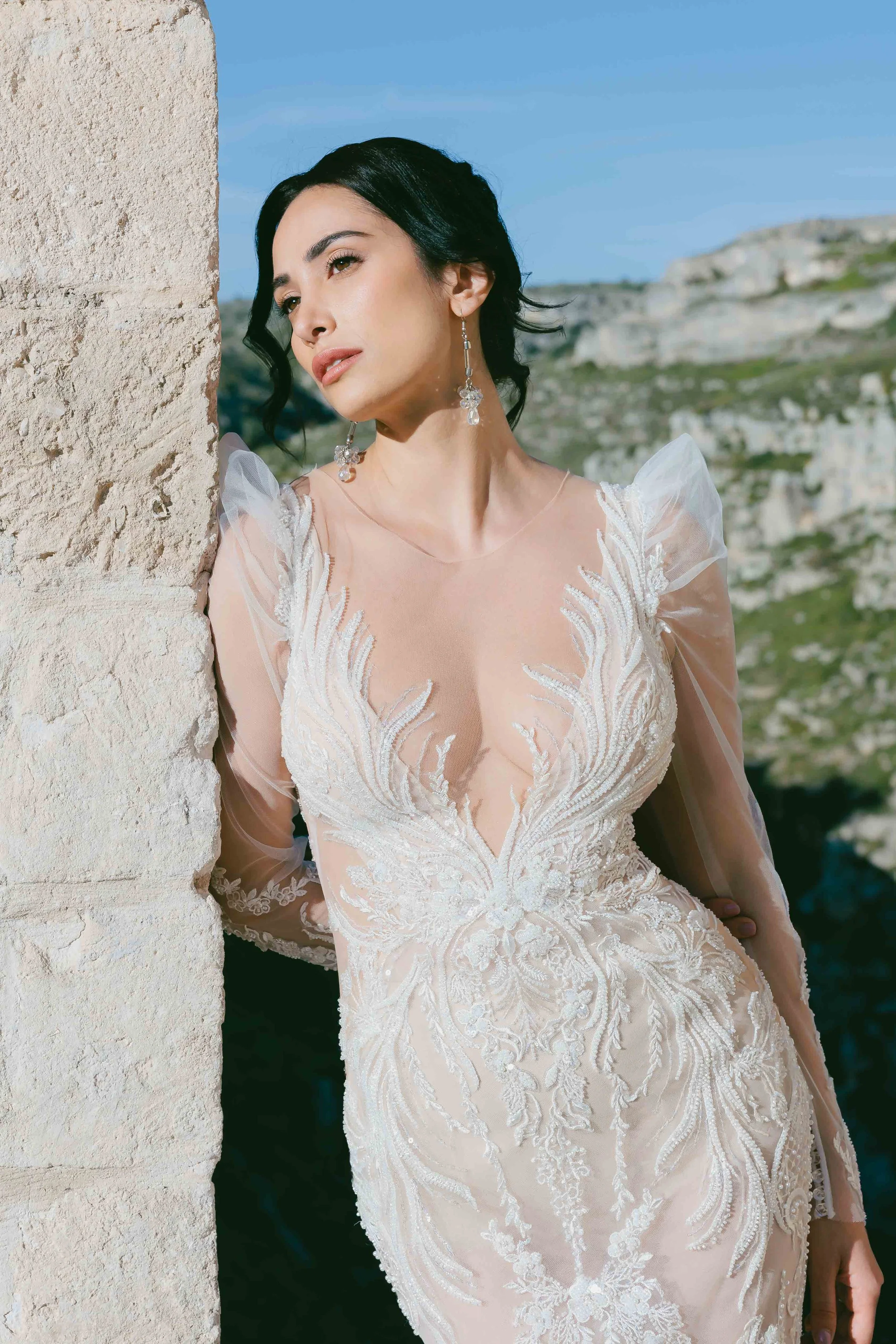 Bride in feather inspired nude and white wedding gown leaning on white limestone wall and looking into bright sun