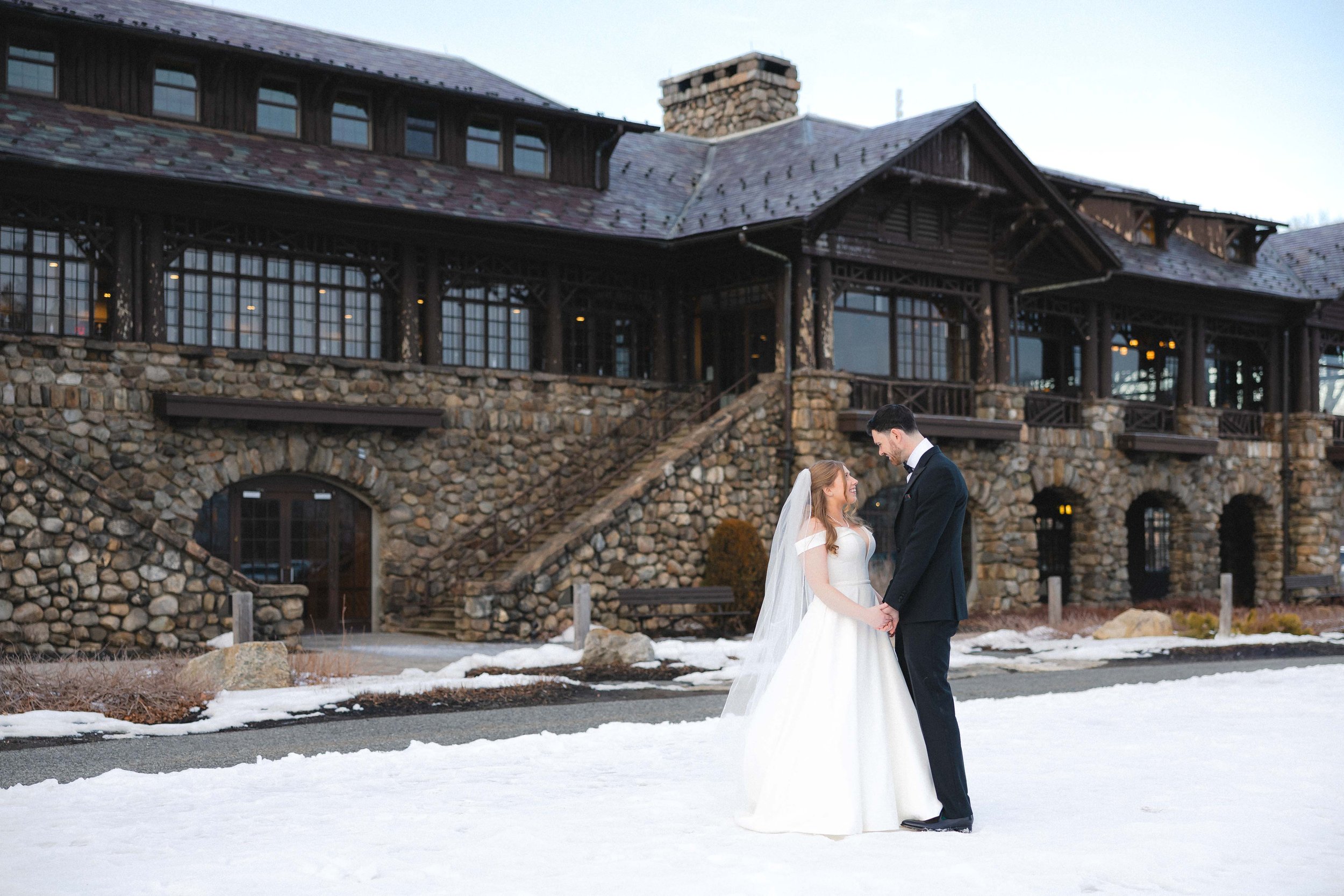 A bride and groom holding hands and smiling at each other outside on snow-covered ground, with a rustic stone and wood lodge building in the background.