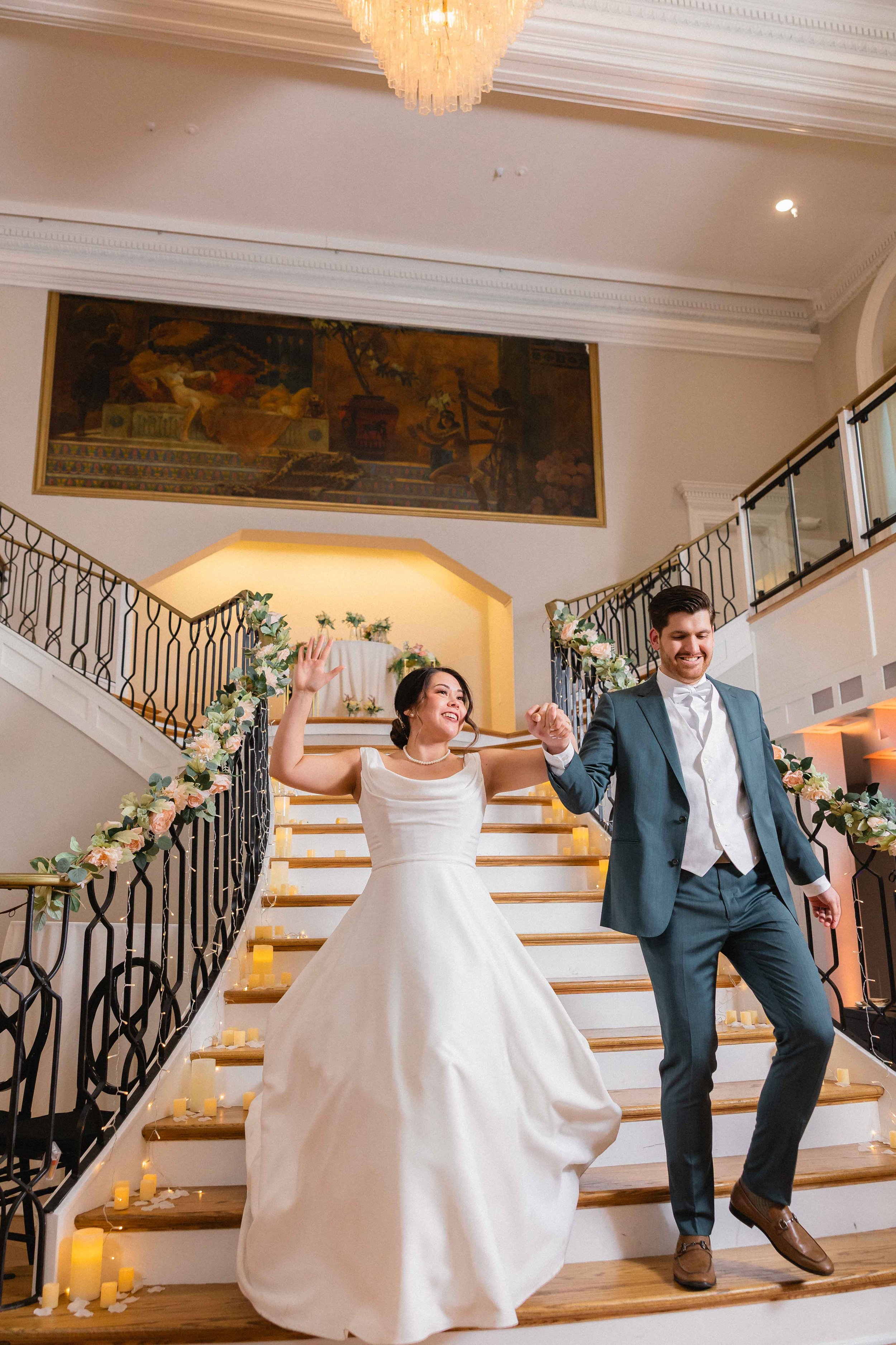 A bride and groom celebrating on a staircase decorated with candles and floral garlands, smiling and holding hands during their wedding.