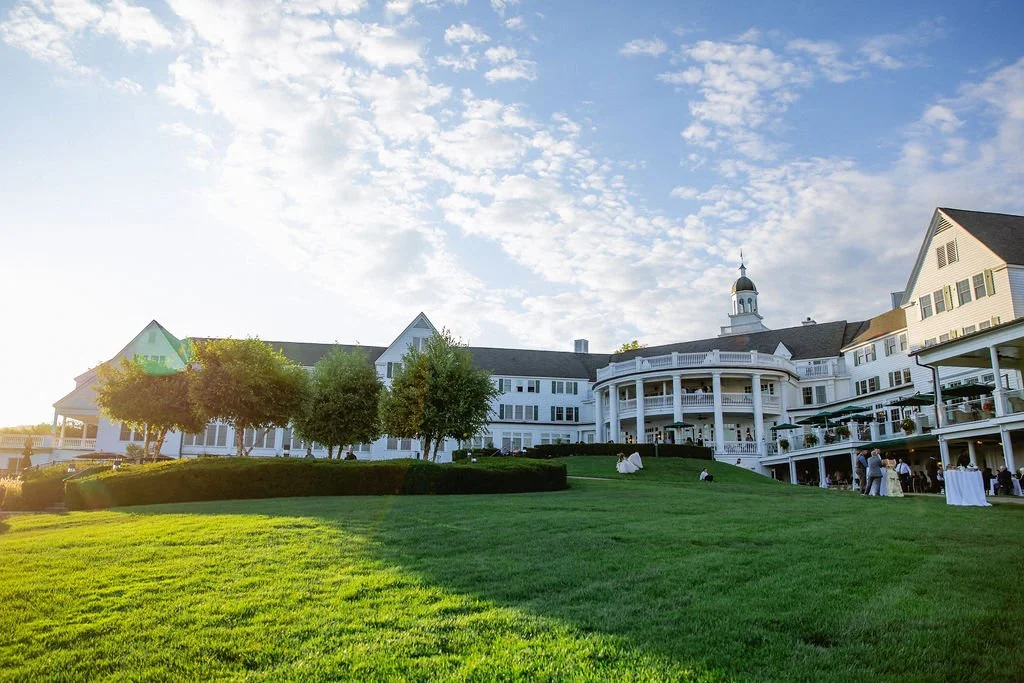 Large white hotel or resort building with a covered porch and balconies, green lawn in foreground, trees, and a partly cloudy sky.