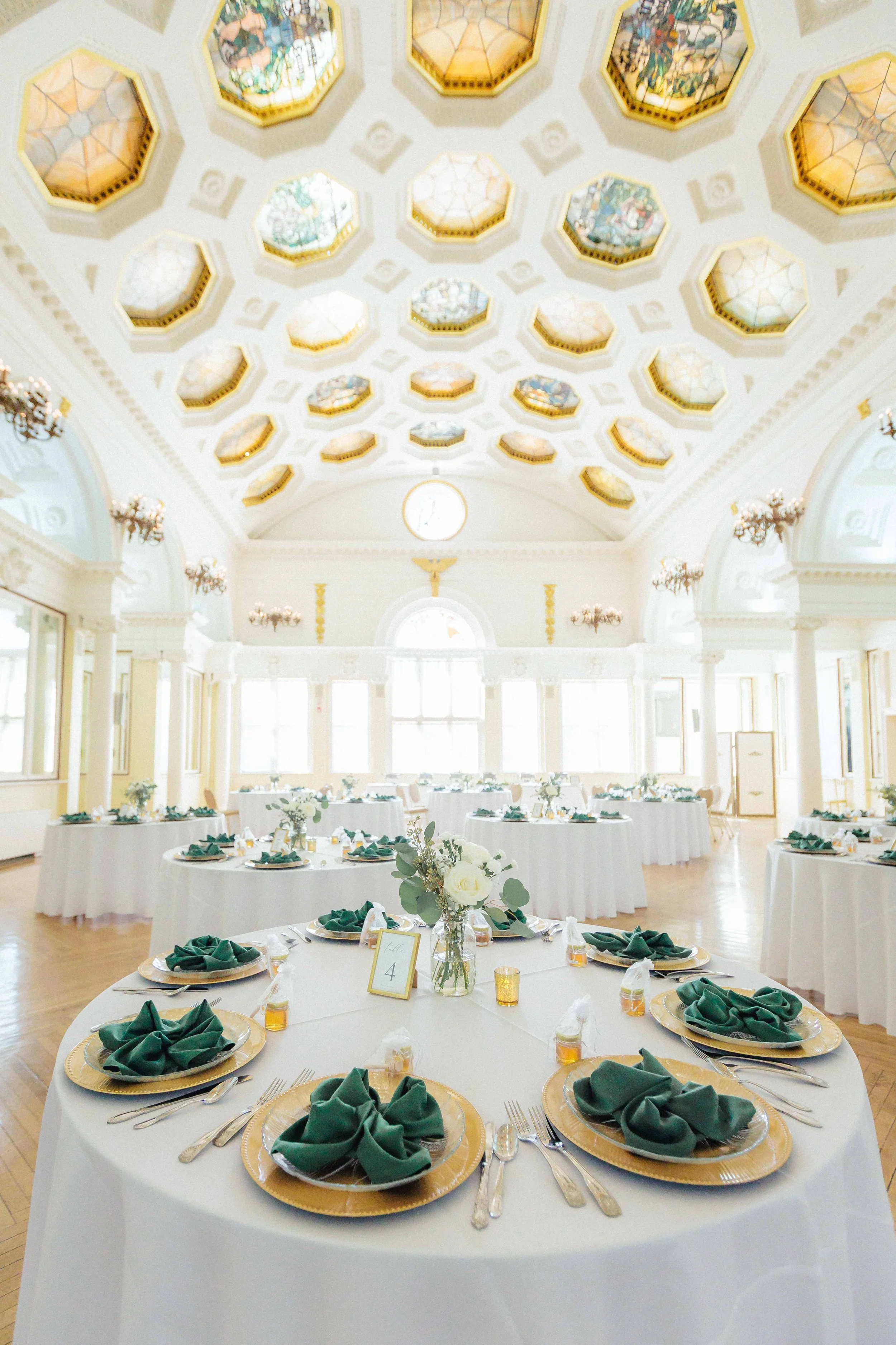 Elegant banquet hall decorated for a wedding reception with round tables set with gold chargers, green napkins, white flowers, and candles, under a decorated ceiling with honeycomb-shaped stained glass panels.