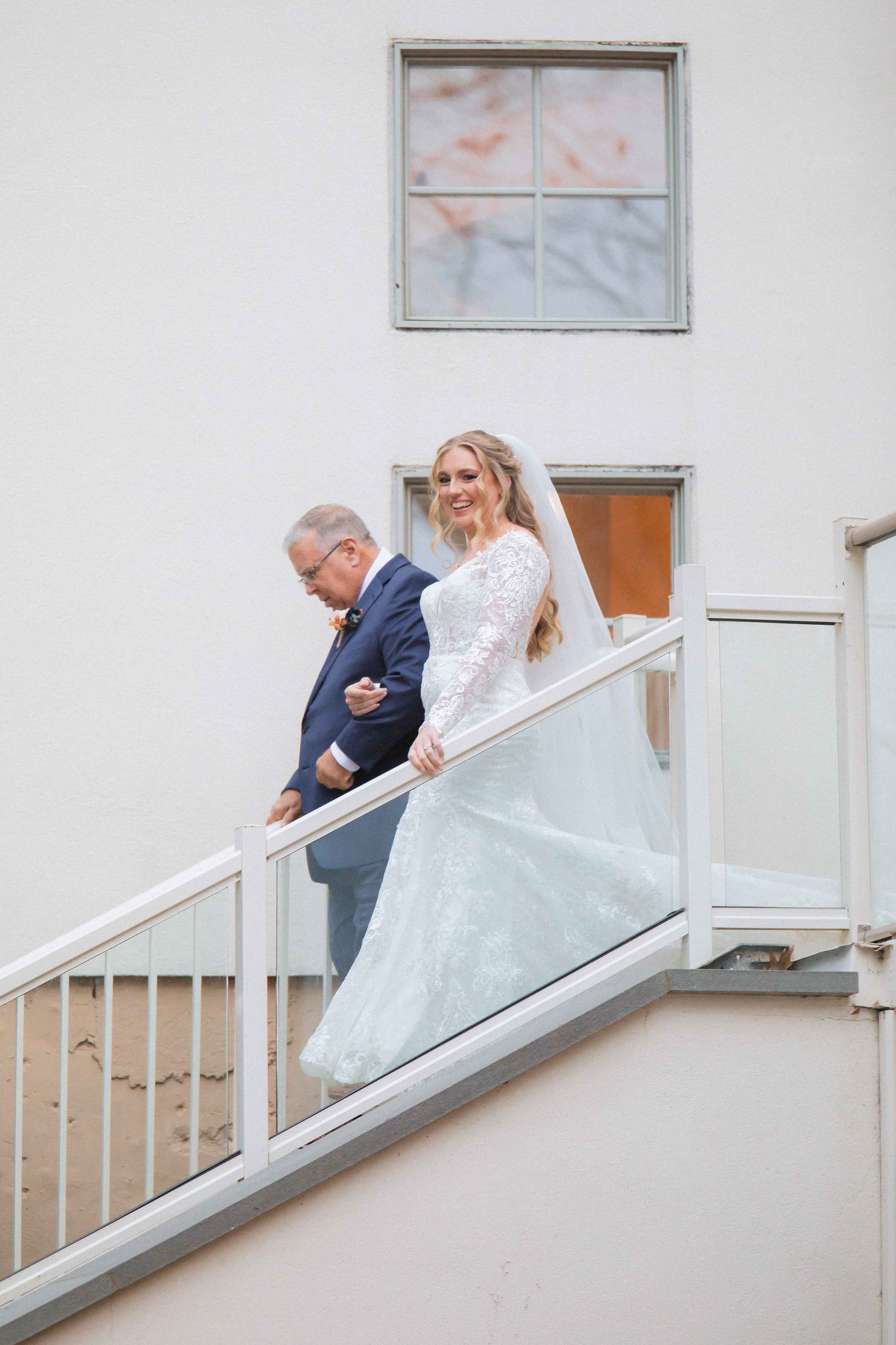smiling bride descends stairs accompanied by her father before the ceremony at the waterview wedding venue in monroe ct