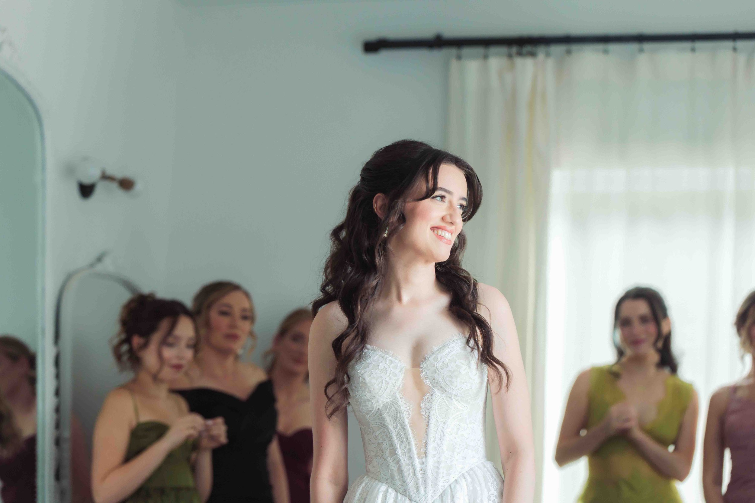 A woman with dark curly hair, wearing a white lace dress, smiling at a gathering of women in elegant dresses in a bright room with light-colored walls and curtains.