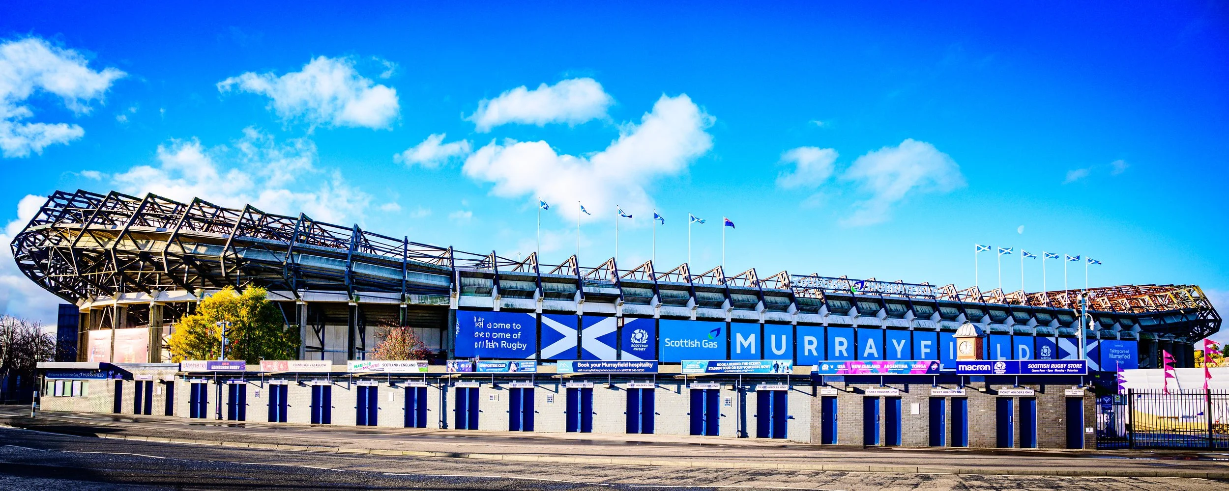 Exterior view of Murrayfield Stadium in Edinburgh, Scotland, with blue sky and Scottish flags flying from the top.