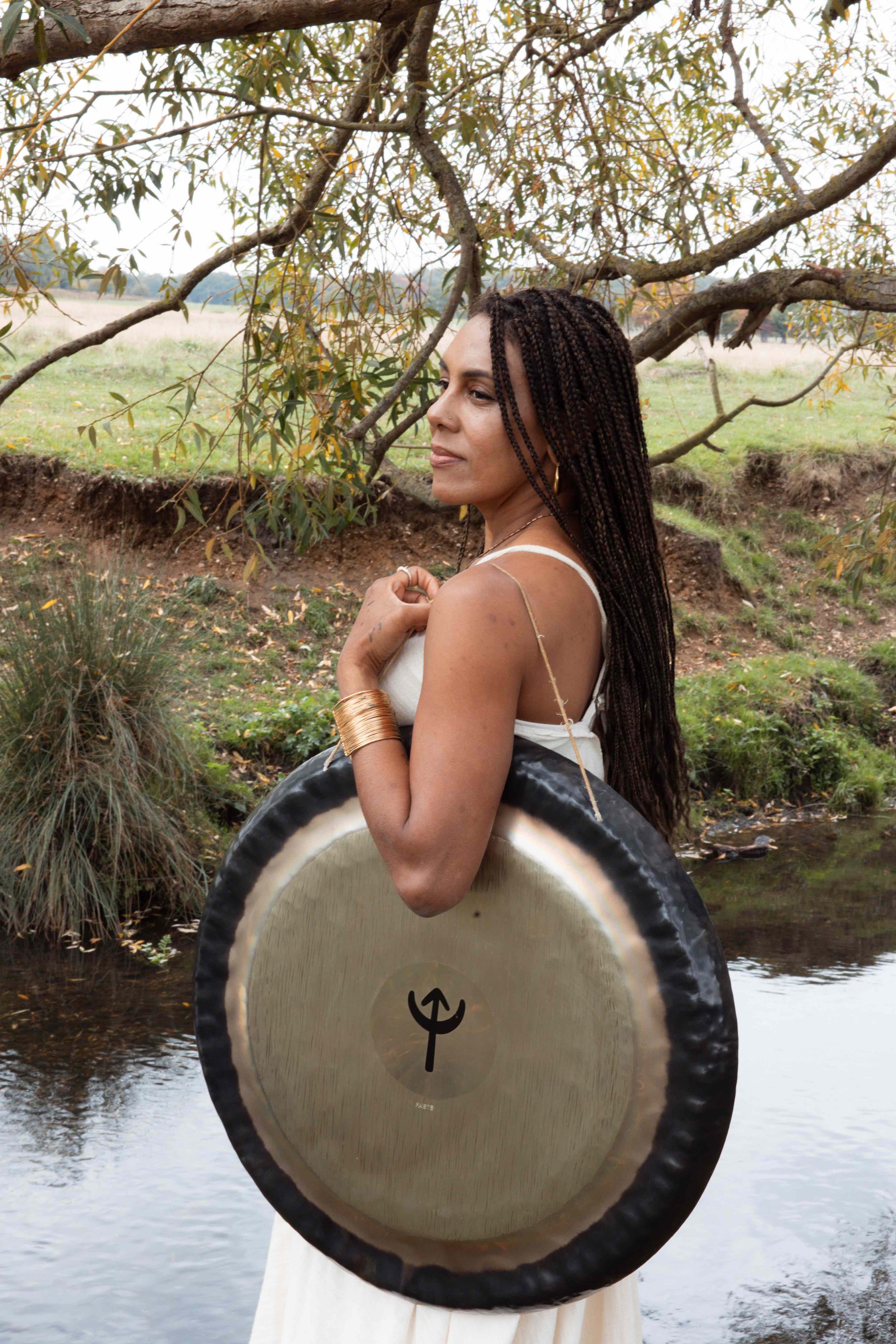 A woman with long braided hair standing beside a stream, holding a large gong with a black rim and a symbol on it, under a tree with green leaves.