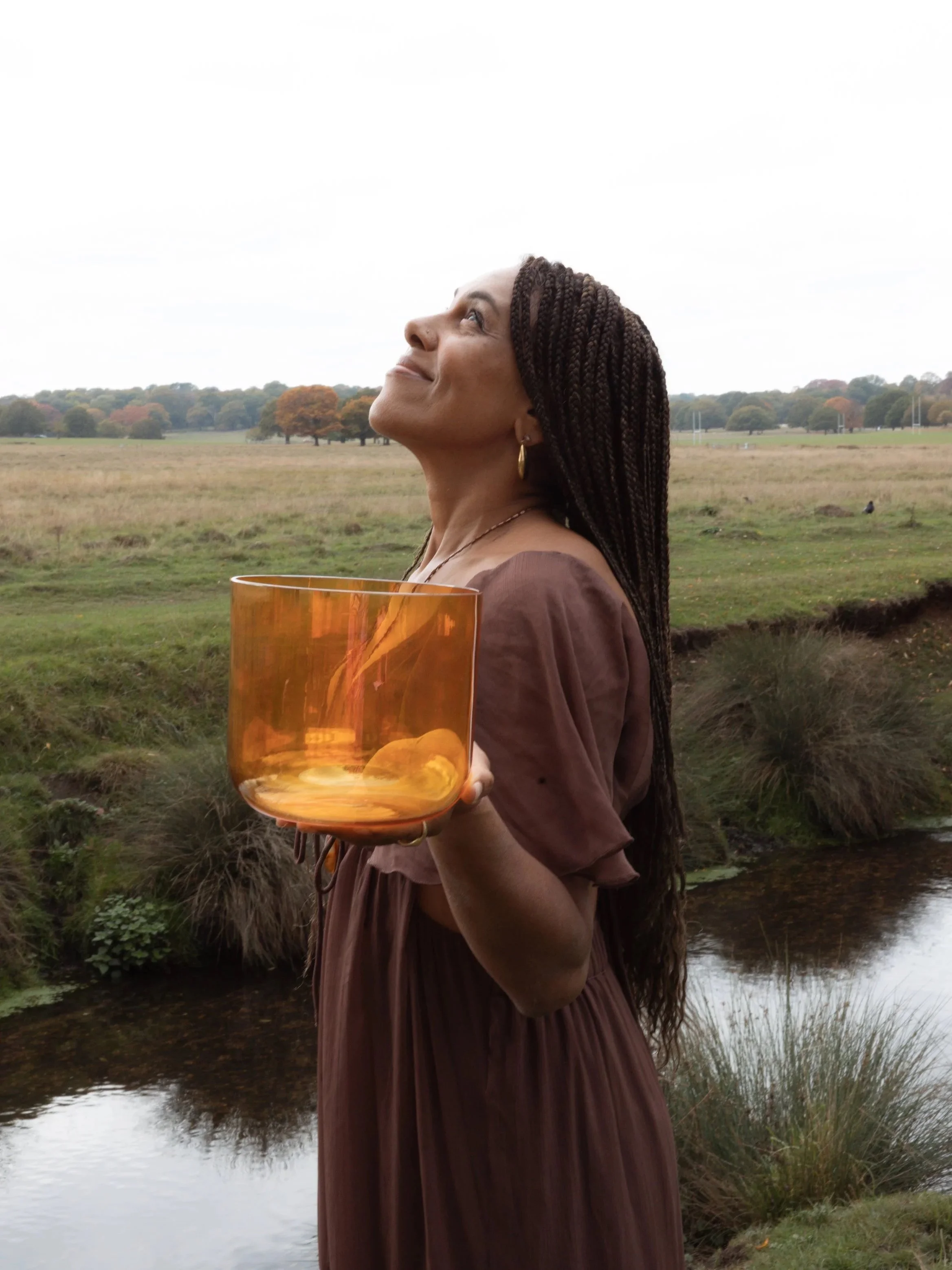A woman with long braided hair holding an orange glass bowl outdoors near a small stream, smiling and looking up in a peaceful setting with grassy field and trees in the background.