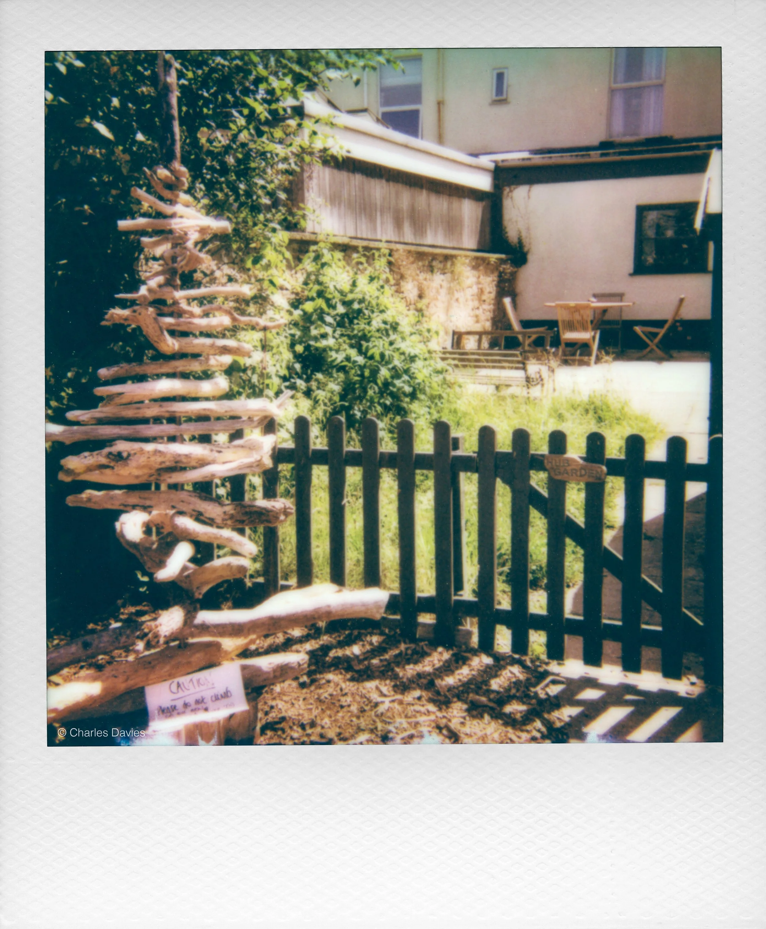 A pile of driftwood arranged vertically in front of a garden fence with green foliage and a house in the background, with warning signs at the bottom.